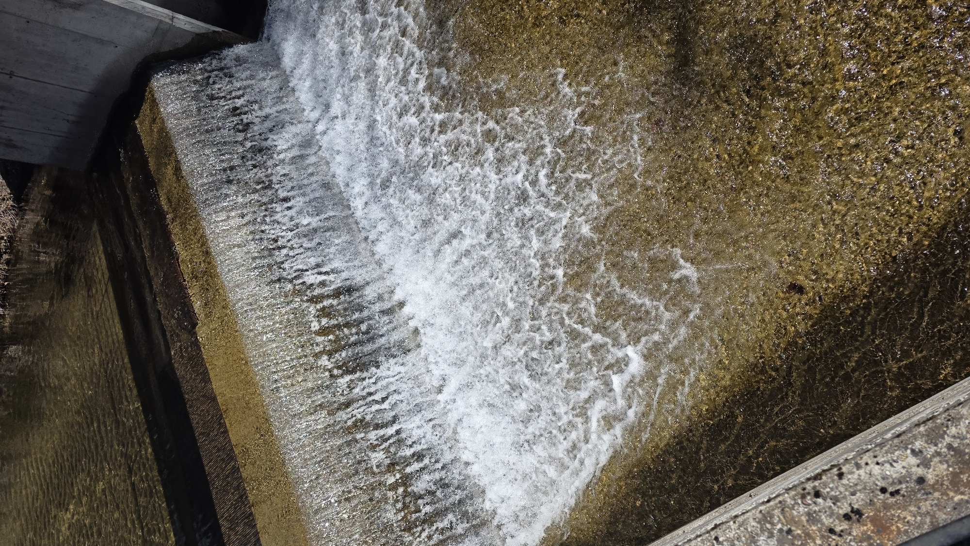 The image depicts a small, artificial waterfall or dam, likely within an urban or park setting. The water cascades over a series of concrete steps, creating a turbulent flow of white water against the calmer, amber-colored water below. The surrounding area appears to be stone or concrete structures, possibly part of the waterfall's construction or landscape design. The image is captured from a slightly elevated, almost overhead perspective, emphasizing the flow of water and the textures of the concrete. The location is mentioned to be in Blagoevgrad, Bulgaria, which may provide context to the specific design or purpose of this small waterfall.
