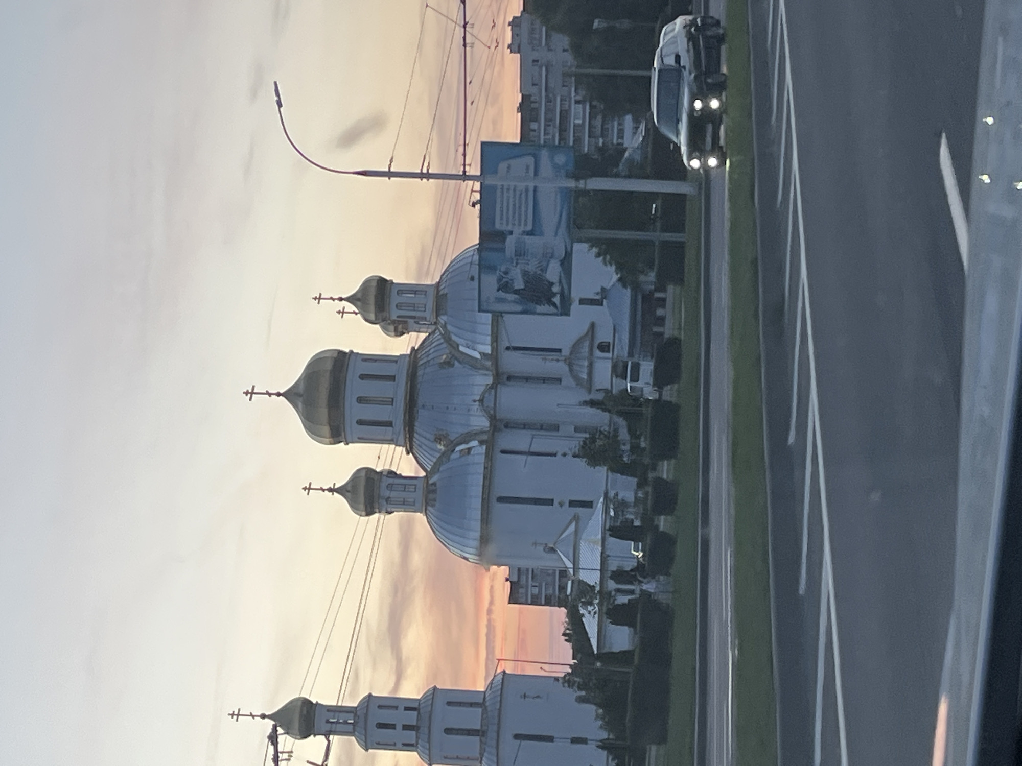 The image depicts a street view in the city of Brest, Belarus, during what appears to be sunset or early dawn. A large, white Orthodox church with multiple golden domes and crosses dominates the background. To the left of the church is a tall, white bell tower. In front of the church, there are trimmed hedges and some low buildings.
A billboard displaying an owl and some text is visible to the right of the church. A street lamp with a curved pole stands near the billboard. In the foreground, a dark-colored SUV with its headlights on is driving on the road. The road in the foreground has white lane markings. Power lines are visible crisscrossing the sky above the church and bell tower. The sky exhibits a gradient of colors, with shades of pink, orange, and light blue, suggesting the time of day. Several residential buildings are visible in the distance behind the church.
