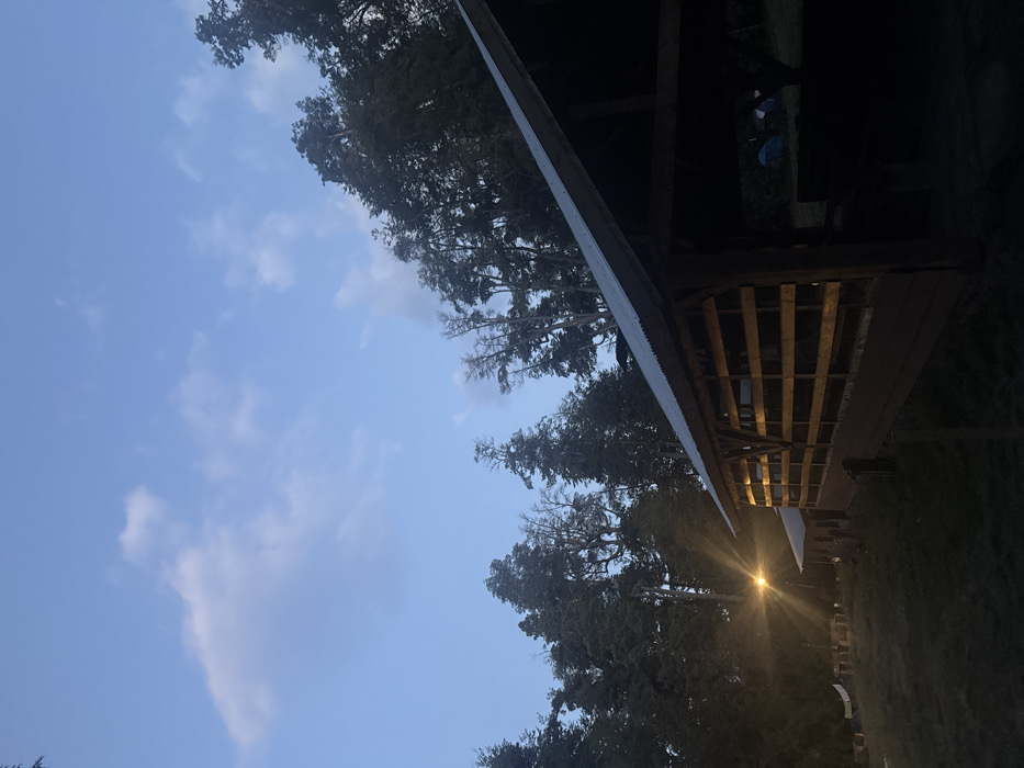 The image displays an outdoor scene at twilight or dawn in Villa La Angostura, Argentina. The upper portion reveals a light blue sky with scattered cumulus clouds, transitioning to a darker tone towards the horizon. Dominating the midground and right foreground is a rustic, open-sided wooden pavilion featuring a corrugated metal roof and exposed wooden supports. Its long side, equipped with horizontal wooden slat railings, is partially illuminated by an unseen light source within. The interior of the pavilion is deeply shadowed, obscuring specific details but suggesting the presence of benches or tables. To the left of the pavilion, a bright artificial light source with a distinct starburst flare illuminates a patch of dark grass. In the background, a dense forest of tall coniferous and deciduous trees forms a dark silhouette against the sky. Within the shadowed forest area, behind the pavilion, the faint outlines of multiple tents, including at least one blue tent, are discernible, indicating a camping or outdoor accommodation site. A clothesline with light-colored items hangs near the artificial light source on the left. No human figures are clearly visible in the frame.