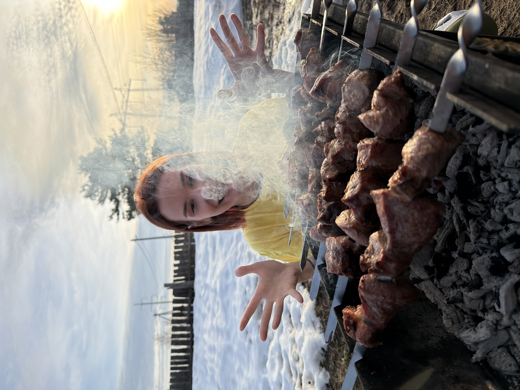 The image depicts an outdoor scene in Chaikovsky, Russia, featuring a barbecue in progress. A metal grill in the immediate foreground holds numerous skewers laden with cubed meat, actively cooking over glowing charcoal embers and emitting a dense plume of white smoke. Behind the grill, a female individual with reddish-brown hair, wearing a yellow t-shirt, is visible from the chest up. She displays a wide smile, and her hands are raised and spread outwards, positioned on either side of the smoke. The ground beneath the grill is dark earth, transitioning to patches of melting snow in the mid-ground. A rustic wooden fence, constructed from vertical logs, forms a boundary behind the snow-covered area. The distant background reveals sparse, bare trees and two prominent electrical utility poles with overhead lines against a partly cloudy sky. The sun, low on the horizon, casts a warm, bright light from the right side of the frame.
