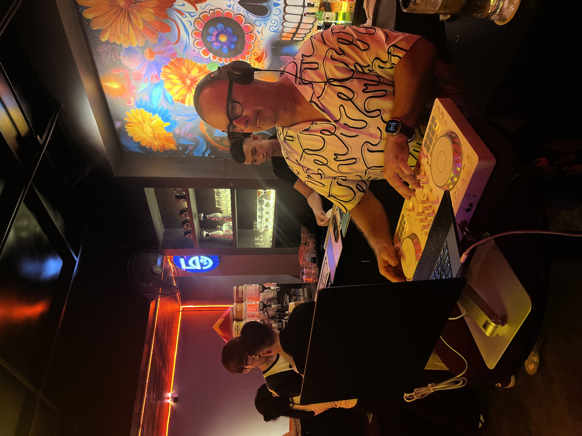 A medium shot depicts a DJ, wearing glasses and headphones, operating a mixing console at a bar in Đà Nẵng, Vietnam. He's wearing a patterned shirt and has a wristwatch visible on his left wrist. The mixing console is positioned near a laptop. Two individuals are partially visible in the background. The bar setting includes a decorative wall with colorful flower patterns, shelving displaying bottles, and a fan. There is also a light and neon sign present in the scene.
