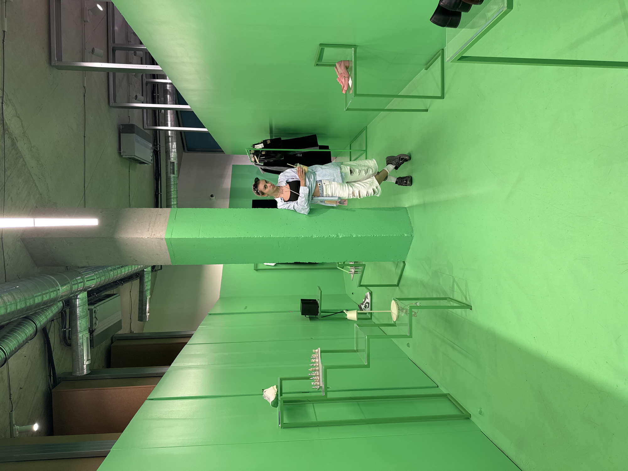 The image shows a person standing inside a retail store in Tbilisi, Georgia, with bright green walls and floor. The person is leaning against a column, holding a drink. The store appears to be a shoe store, with various shoes displayed on green metal shelves. There is also clothing on a rack in the background. The ceiling has exposed industrial pipes and lighting fixtures. The overall impression is modern and minimalist, with a bold color palette.