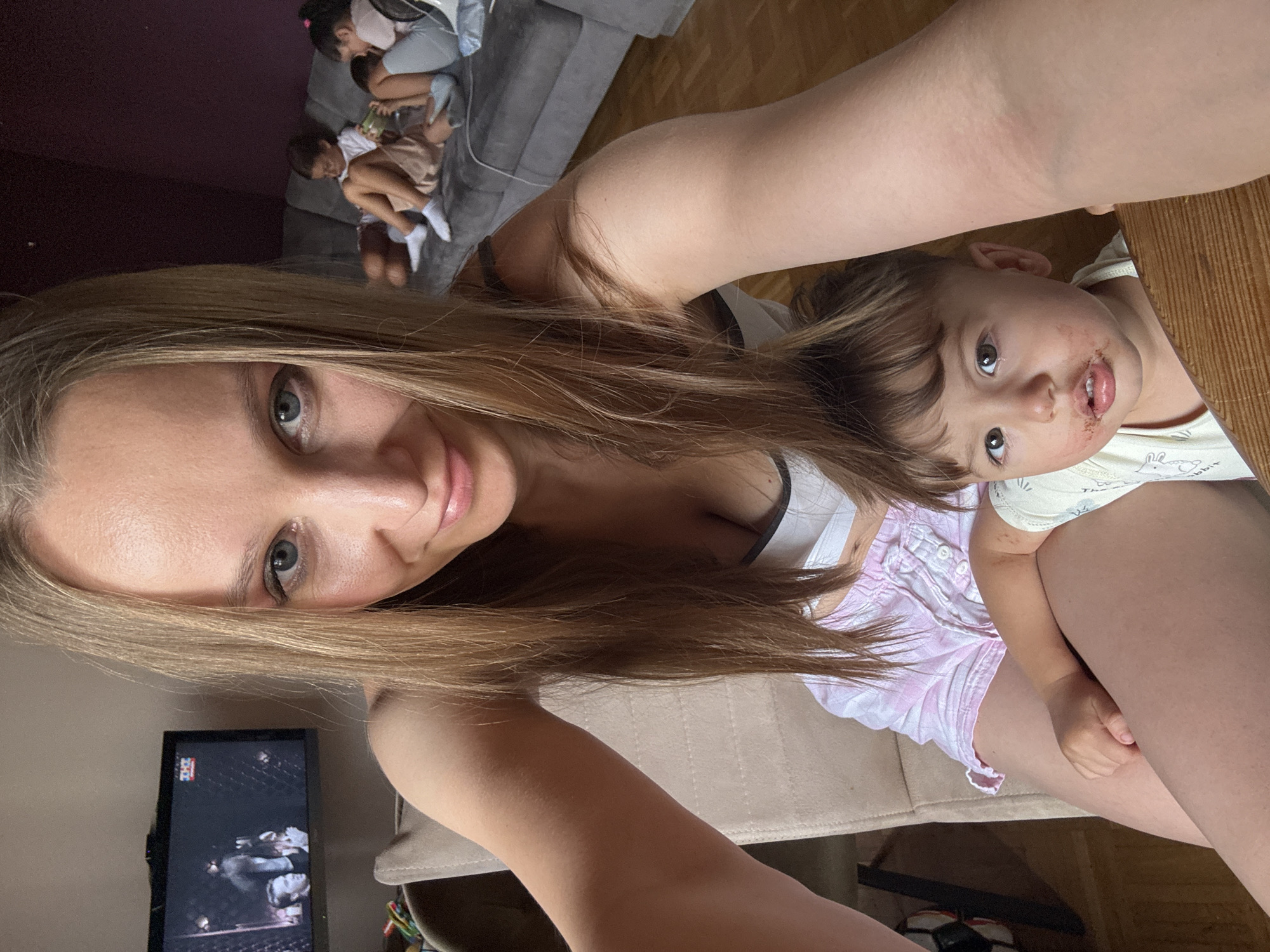In a domestic indoor environment, likely a living room, a woman is taking a selfie. She is seated, with a young child positioned on her lap. The woman has long blonde hair and is wearing a white and pink top. The child has brown hair and is wearing a light-colored outfit with a rabbit pattern. The child appears to have chocolate around their mouth. In the background, other children are visible on a sofa, and a television is mounted on the wall. The overall scene suggests a casual family moment. The location is identified as Überherrn, Germany.