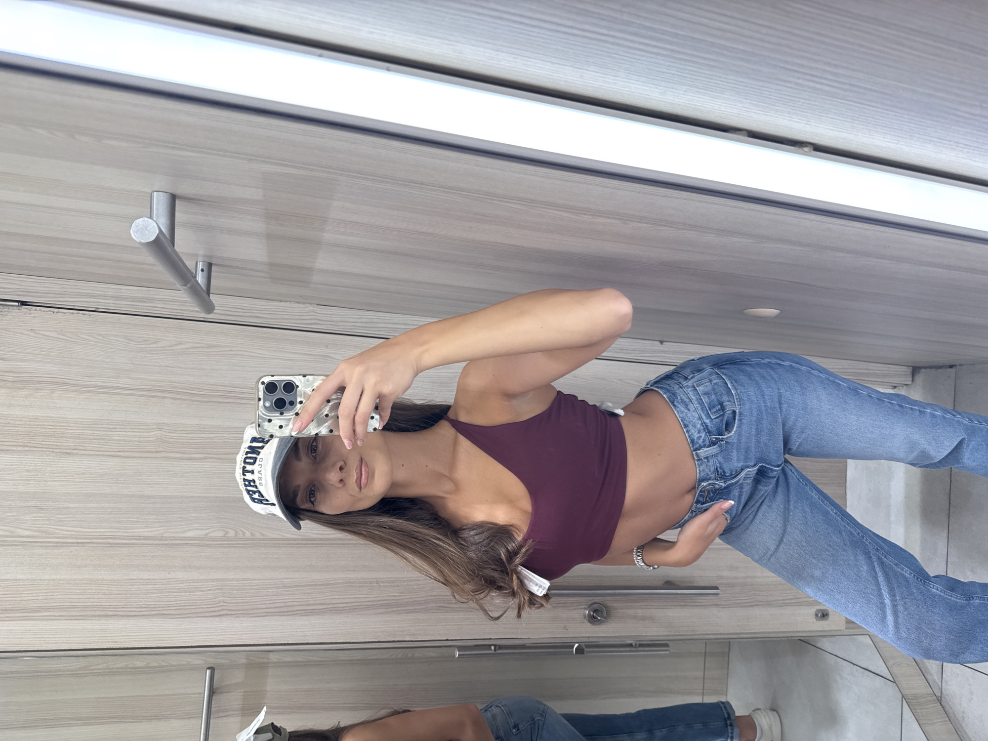 A young woman is taking a mirror selfie in what appears to be a dressing room. She is wearing a maroon tank top, light blue jeans, and a baseball cap. She is holding her phone in her right hand to take the picture. The dressing room has wooden paneling and silver hardware. The location is not discernible as Burgas, Bulgaria, from the image.