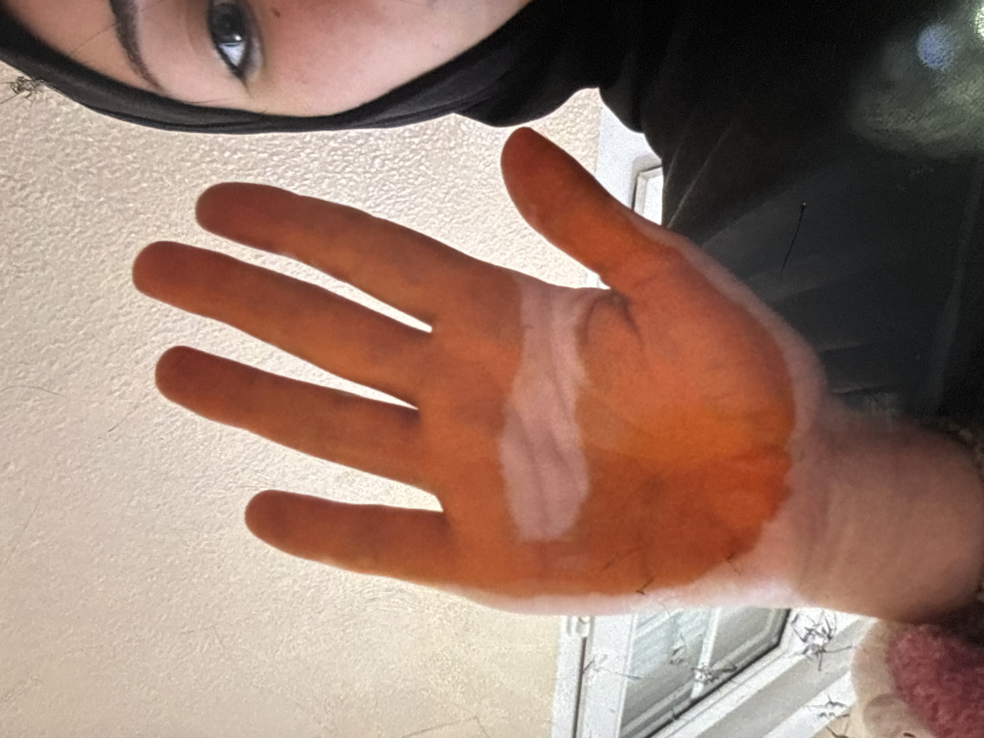The image shows a close-up, low-angle view of a person's hand, which appears to be stained with an orange substance on the palm and fingers. The person is wearing a black hijab, and only a portion of their face, including an eye and eyebrow, is visible on the right side of the frame. The background consists of a textured, light-colored ceiling and a window with visible panes and mullions. There are no discernible features in the image that would indicate a specific location within the city of Orihuela, Spain. The image appears to be a selfie, with the hand held up towards the camera.