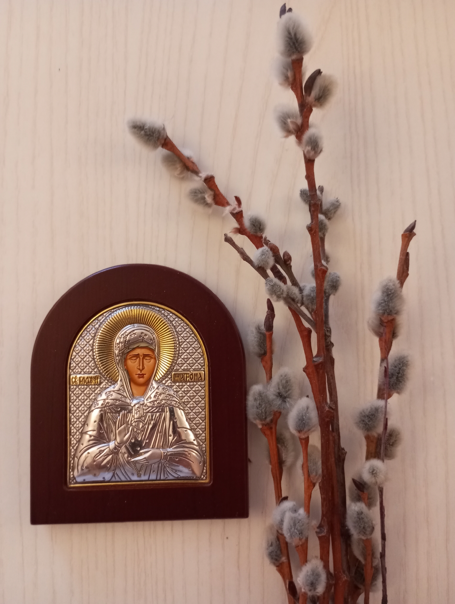 The image shows a religious icon and pussy willow branches placed against a light-colored, possibly wooden, surface. The icon is in a dark wood frame with an arched top. The icon itself is a silver-colored image of a woman with a golden halo, likely a religious figure. To the right of the icon are several branches of pussy willow, with their characteristic soft, furry buds. While the image does not explicitly confirm the location, the user mentions Chaikovsky, Russia. The context suggests the image may have a religious or cultural significance, possibly related to Easter celebrations, as pussy willows are often used in place of palms in some Eastern Christian traditions.