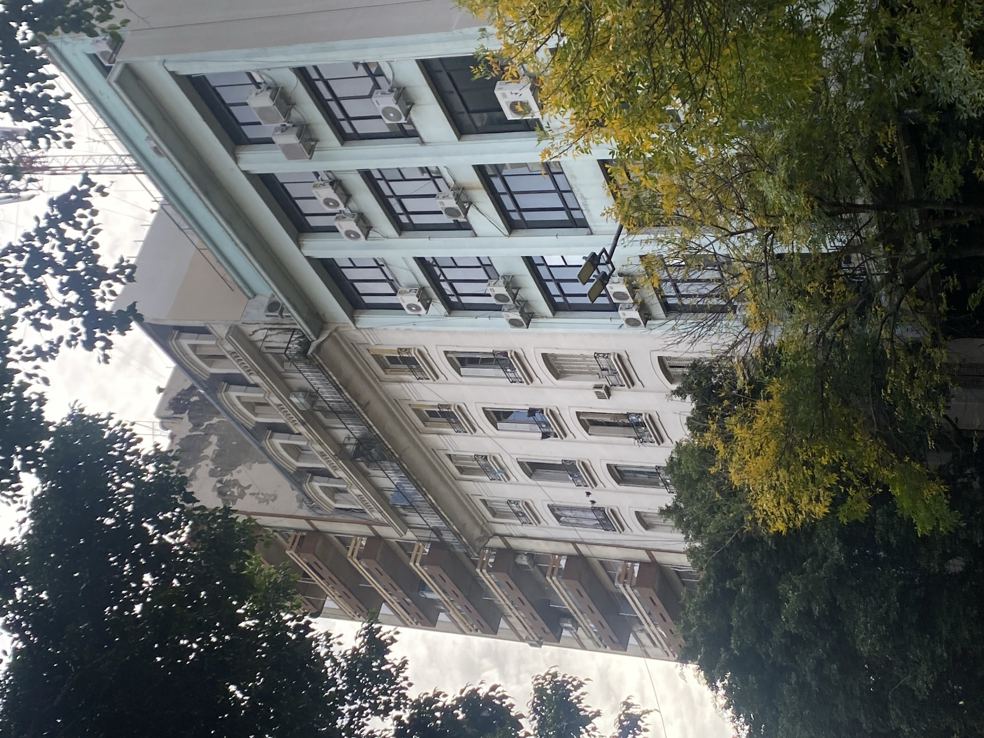 The image shows a tall, white building with multiple stories in Buenos Aires, Argentina. The architecture suggests an older building. There are trees obscuring parts of the facade, implying the building is in an area with some greenery. The building has many windows, some with air conditioning units installed. Some floors have balconies. A streetlight is visible near the top of the building. The sky is overcast. Construction is visible in the upper left corner.