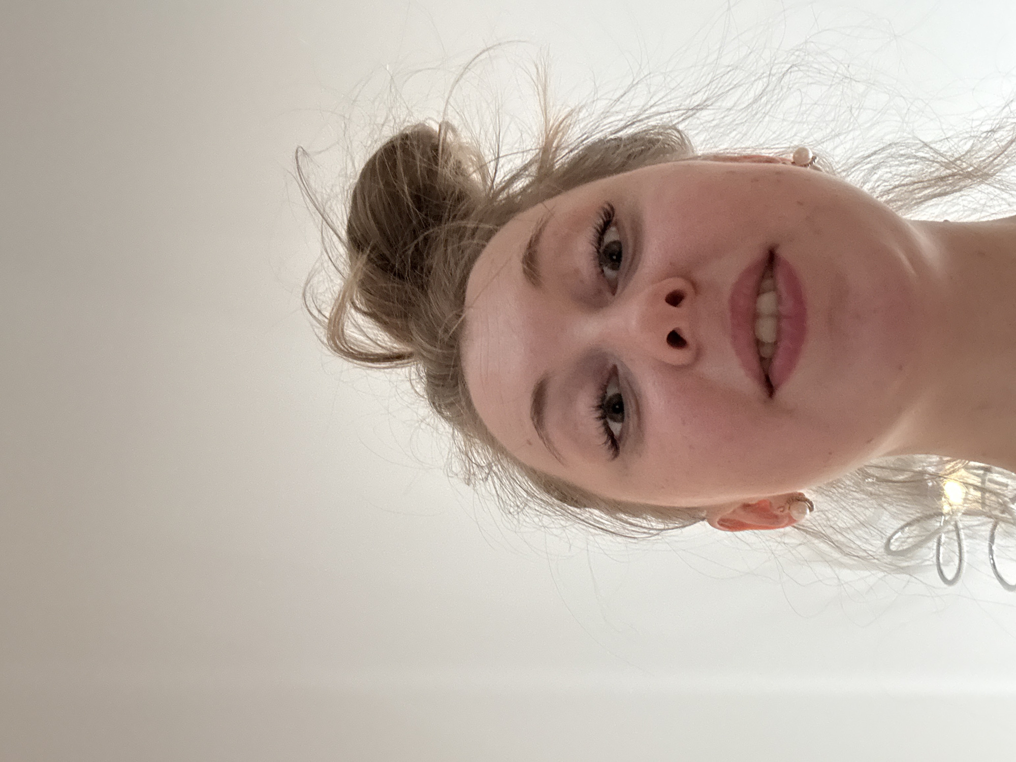 A young Caucasian woman with blonde hair styled in a high bun is positioned in the upper-right quadrant of the frame, looking down at the camera from an elevated perspective. She has fair skin, prominent cheekbones, dark eyebrows, and visible eyelashes. Her mouth is slightly open, revealing her teeth. She wears pearl earrings. The background is a smooth, light beige or off-white color, indicative of a wall or ceiling. The location is Unknown, Unknown.