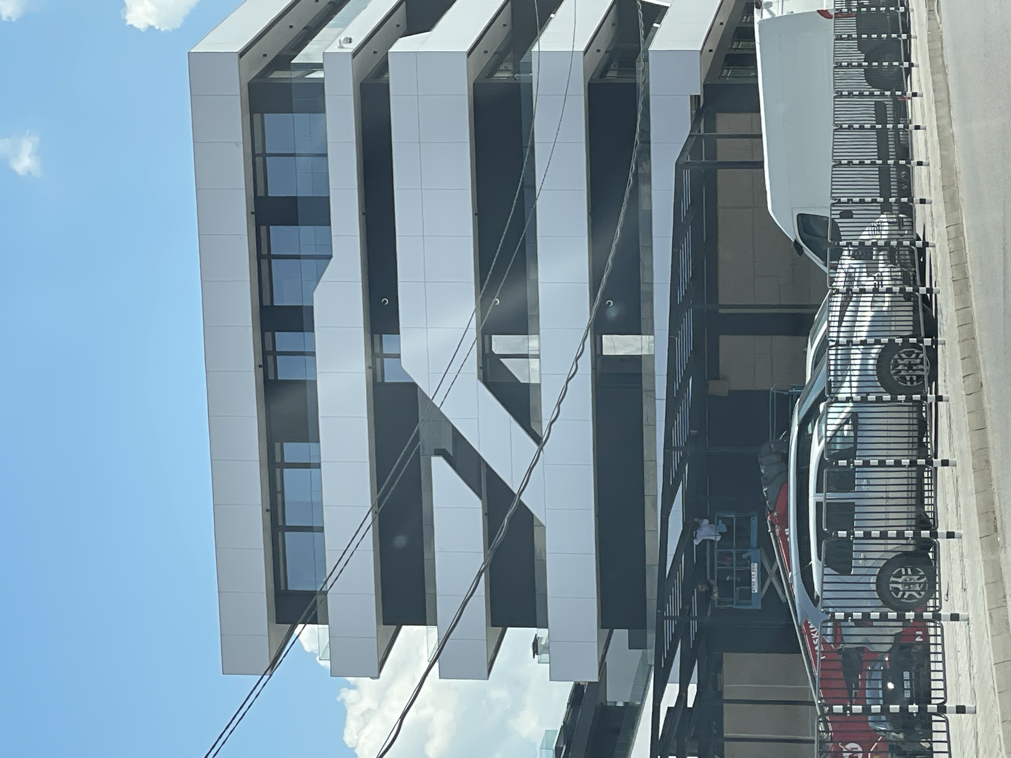 A modern, multi-story building with a black and white facade is under construction. The building features horizontal bands of white paneling interspersed with dark sections and rows of windows. Several balconies with glass railings are visible. In front of the building, there is a black metal fence. Parked behind the fence are several vehicles, including a silver SUV, a red construction vehicle with a boom lift, and a white van. On the boom lift, two individuals appear to be working on the underside of a dark-colored awning structure that extends from the building. Overhead, several dark wires are strung across the scene, obscuring parts of the building. The sky is blue with scattered white clouds. The street in front of the fence is paved. The building is located in Sofia, Bulgaria, on a clear, sunny day.