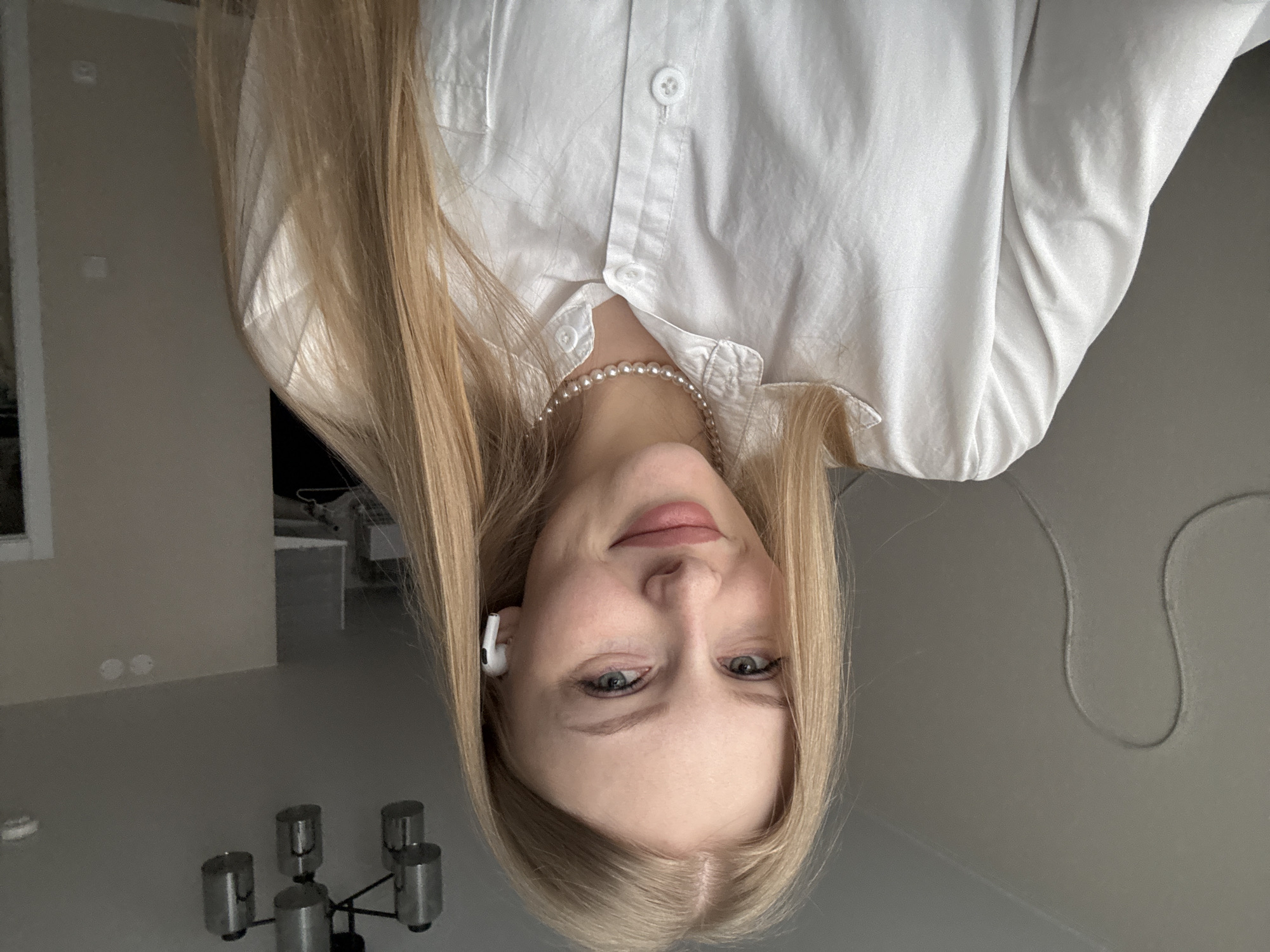 A young woman with blonde hair is wearing a white button-down shirt and a pearl necklace. She has earbuds in her ear. The location is likely an interior space, judging by the presence of a lamp and furniture in the background. There's no information in the image that ties it to Perm, Russia.