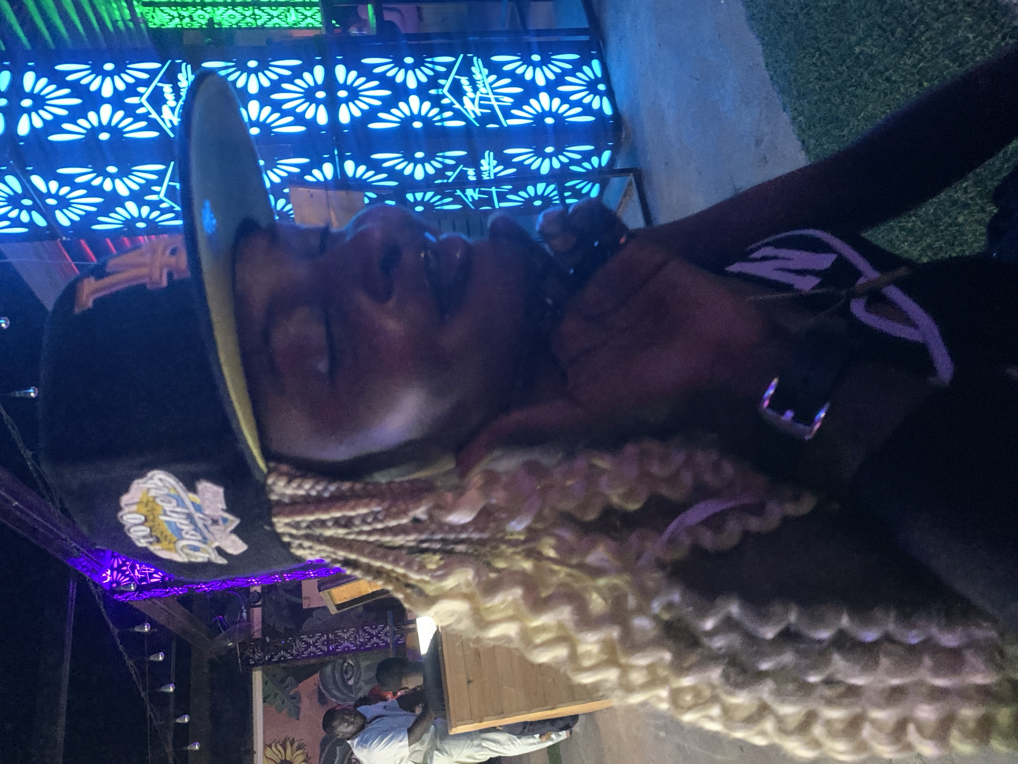 The image shows a medium shot of a Black woman wearing a black baseball cap with a "Dodgers" logo, and blonde braided hair. She is in profile, looking to her right. She is wearing a black top with a white and purple graphic design. She has a necklace on. In the background, there is a brightly lit blue neon sign with a floral design and the text "Road House". To the left, there are people standing near a wooden structure. The environment appears to be an outdoor venue with artificial lighting and what looks like artificial grass on the ground. The location is described as Howayi, Nigeria.