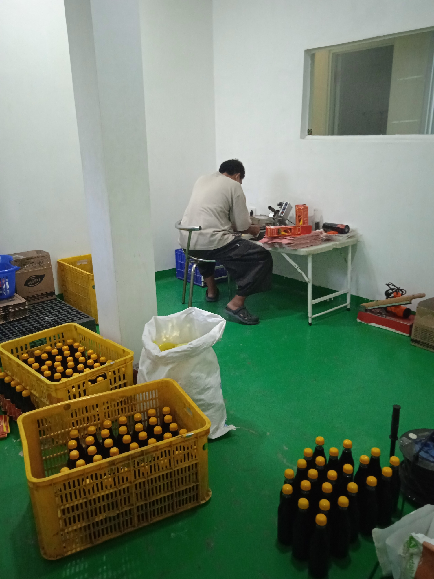 The image depicts an indoor scene, likely a production or storage area in Cileunyi, Indonesia. A person is seated at a table, appearing to be engaged in some form of work involving packaging materials and possibly a sealing machine. Yellow plastic crates filled with dark bottles capped with yellow lids are prominently displayed in the foreground and mid-ground, suggesting a product ready for distribution or storage. A large white bag, possibly containing more product or packaging supplies, is also visible. The floor is a vibrant green. In the background, a small window is set into a white wall, and various boxes and equipment are scattered around the room. The overall impression is one of a functional workspace with a focus on product handling.