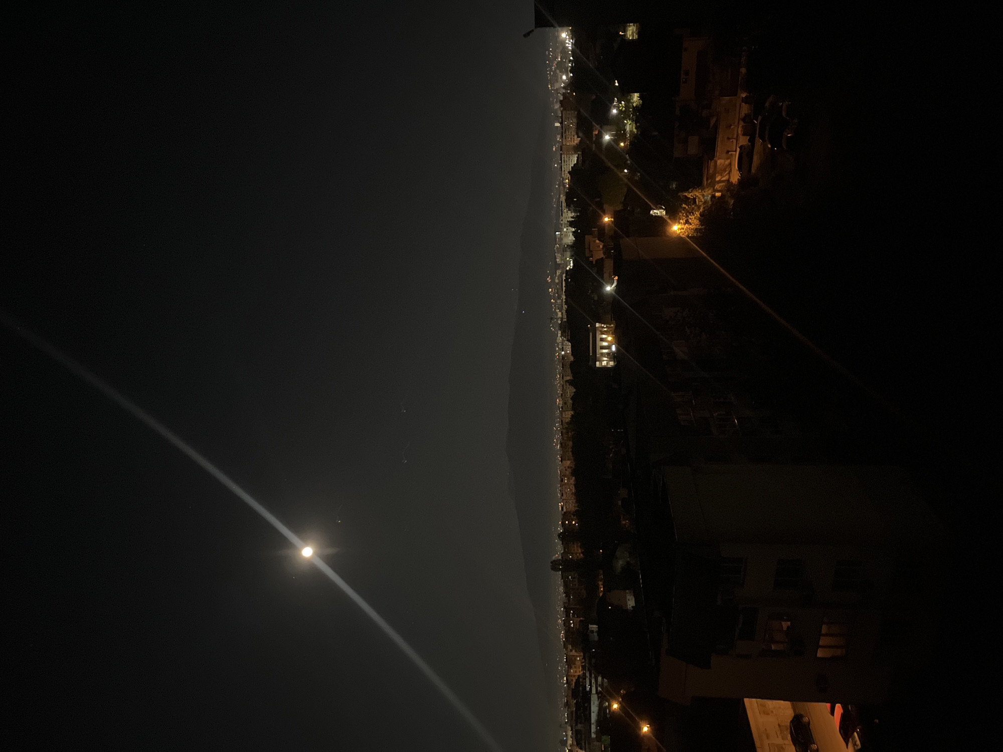 The image shows a nighttime view of Sofia, Bulgaria. The sky is dark with the moon visible in the lower left corner, casting a faint beam of light across the scene. The city lights stretch along the horizon, with darker mountain shapes in the distance. Buildings, streets, and illuminated windows are visible in the right portion of the image. The photo is taken from an elevated position, likely a rooftop or high floor of a building.