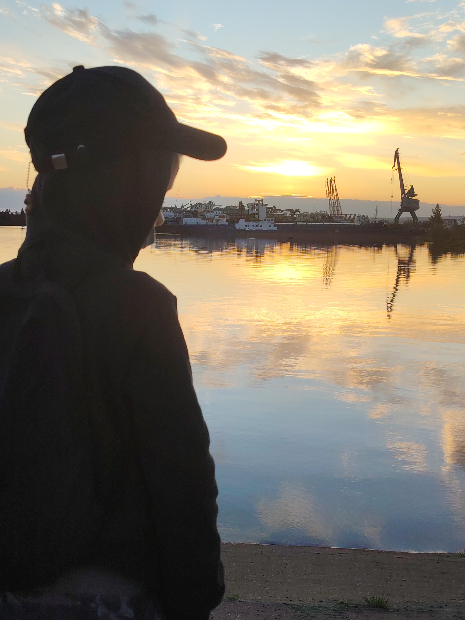 An outdoor, full-frame, eye-level shot shows a person wearing a black baseball cap and jacket, seen from behind and in silhouette. The person is looking out at a body of water. The sun is setting, casting a golden glow across the sky and reflecting on the water. In the distance, across the water, there is a port with several ships and cranes visible. The foreground consists of a sandy or gravelly shore. The overall scene depicts a tranquil evening at a port. The location is the Chaikovsky port on the Kama River in Chaikovsky, Russia, as indicated by historical context and visual cues typical of Russian ports.