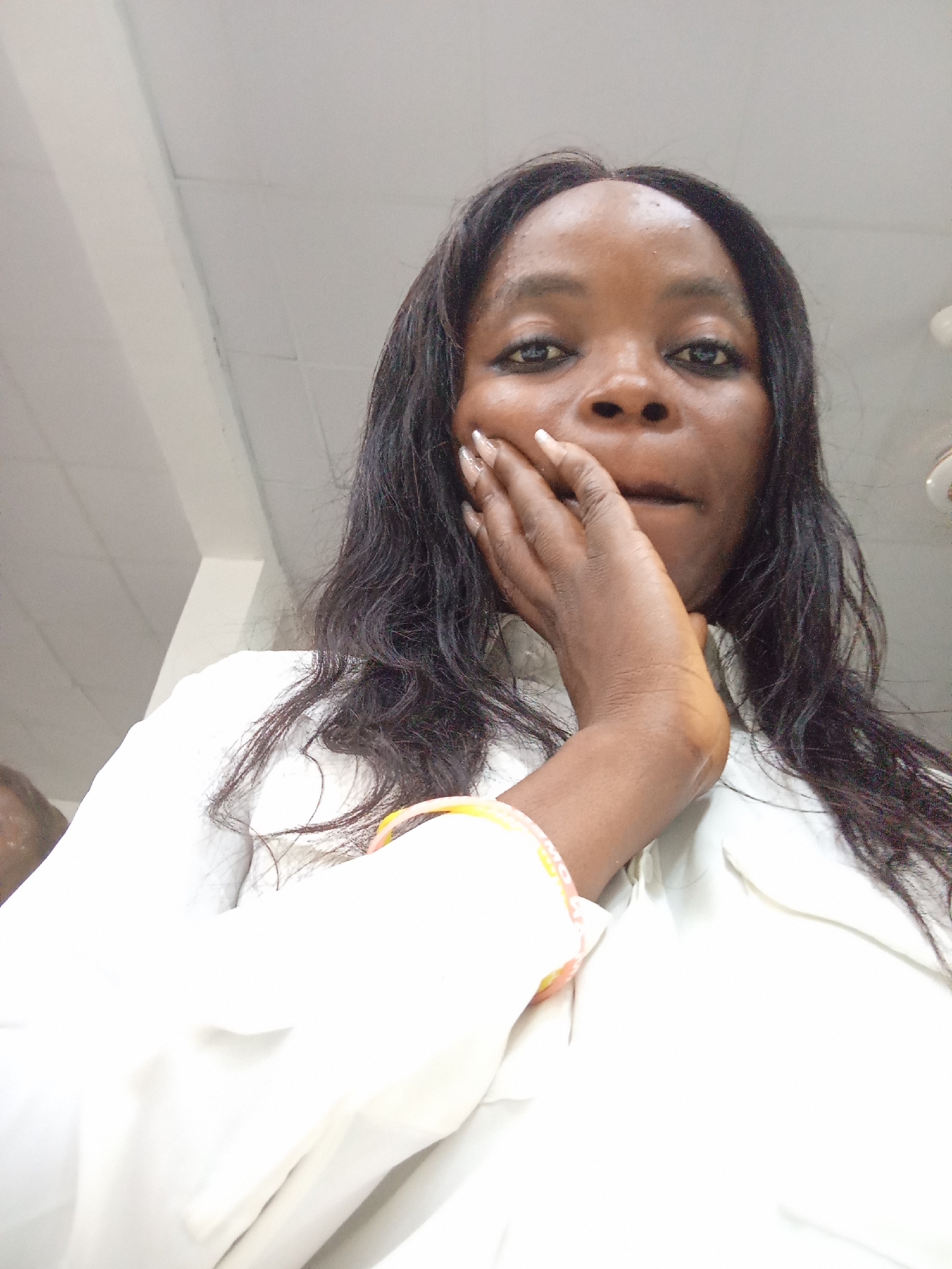 A medium shot is provided of a woman of African descent looking at the camera, resting her chin on her hand. The woman has long, dark hair and is wearing a white collared shirt. She has a bracelet on her left wrist with orange and white beads. The background is out of focus and appears to be an indoor setting with a white ceiling and a partition or wall. The image is taken from a low angle, looking up at the woman. The scene is in Kason, Nigeria.