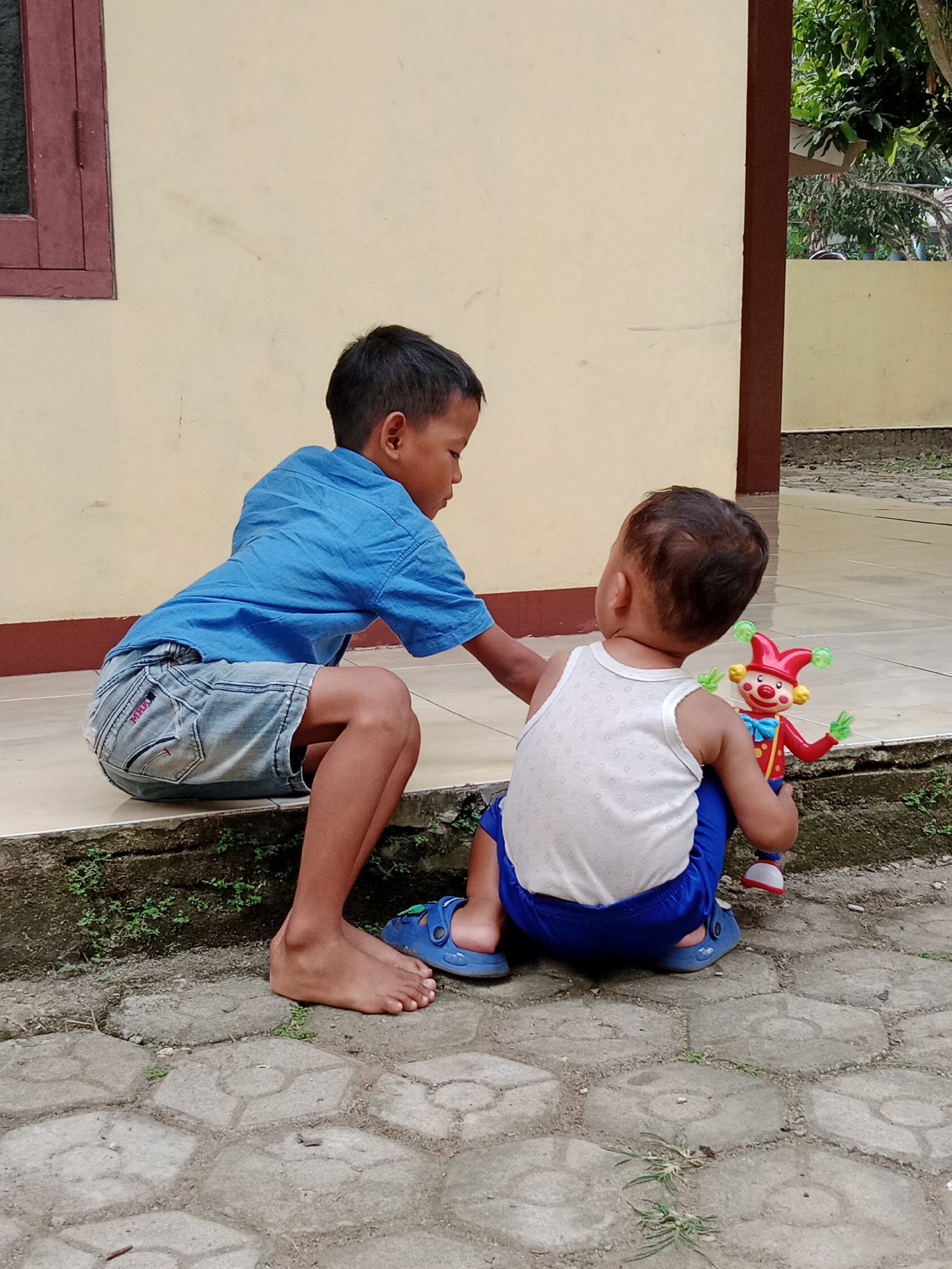 Two young boys are on a paved outdoor area in front of a light yellow building. The boy on the left, appearing to be older, is wearing a blue short-sleeved shirt and denim shorts. He is sitting with his legs bent and his body angled towards the right. He is reaching out his right hand towards the boy on the right. The younger boy on the right is wearing a white tank top and blue shorts. He is sitting and holding a red and green clown toy. Both boys are barefoot except for the younger boy, who is wearing blue sandals. The ground is paved with hexagonal grey tiles, and there is some sparse green vegetation growing between the tiles and along a raised concrete border. A portion of a brown-colored pillar is visible to the right of the building, and behind it, green foliage from trees can be seen. The building has a dark red-framed window on the left side. The scene appears to be in a residential area in Parage, Indonesia, given the architectural style and vegetation.