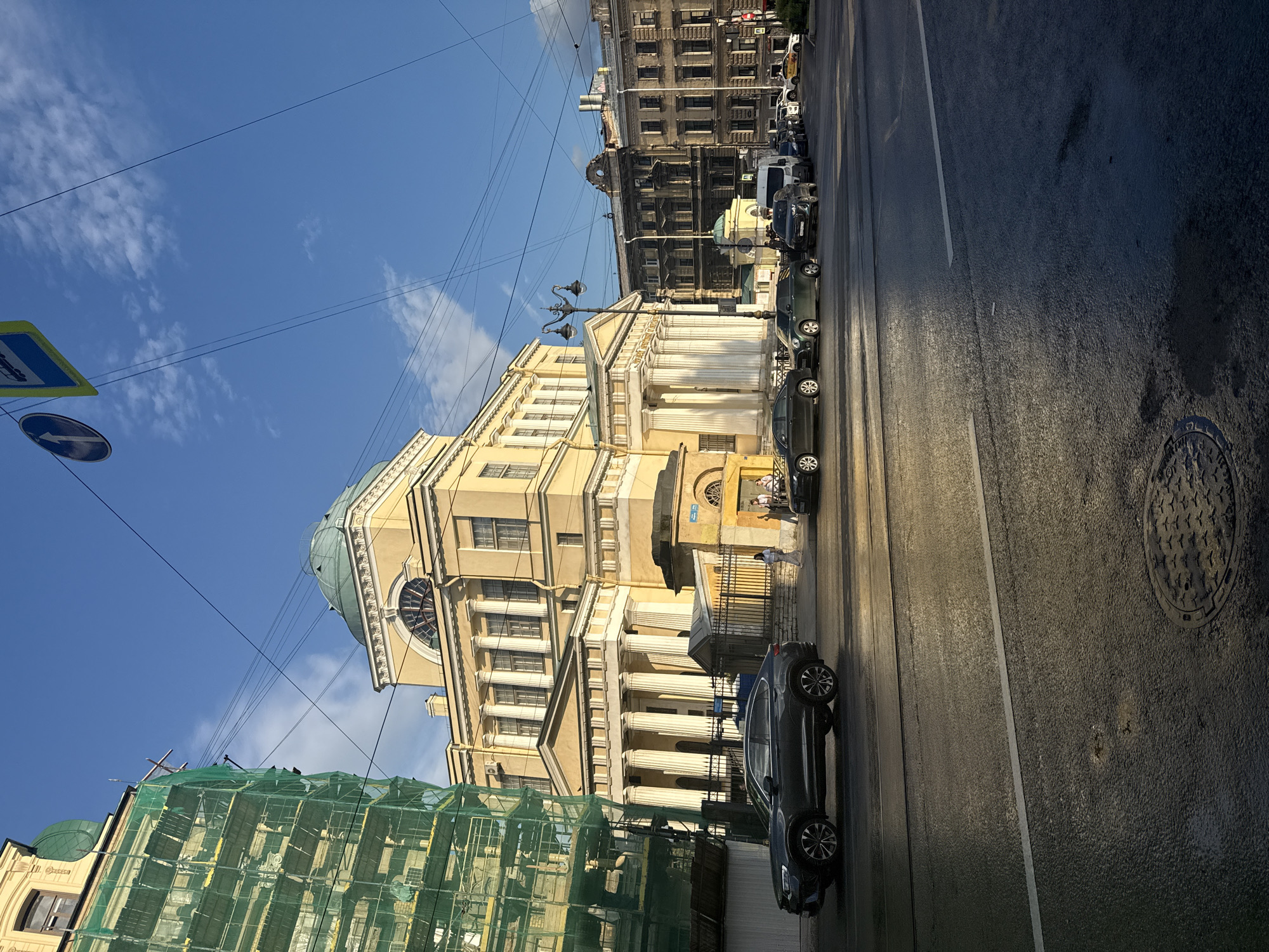 In Saint Petersburg, Russia, on a clear day with scattered clouds, a street scene unfolds. The focal point is a grand, pale yellow building with a prominent dome, featuring classical architecture with columns and arched windows. To the left, a building is partially obscured by green scaffolding and netting, suggesting ongoing renovation or construction. Several cars are parked and driving on the wet asphalt road, reflecting the sunlight. Pedestrians are visible on the sidewalk near the yellow building. Overhead, a network of power lines crisscrosses the sky, and street signs are mounted on poles. The scene captures a blend of historical architecture and modern urban activity.