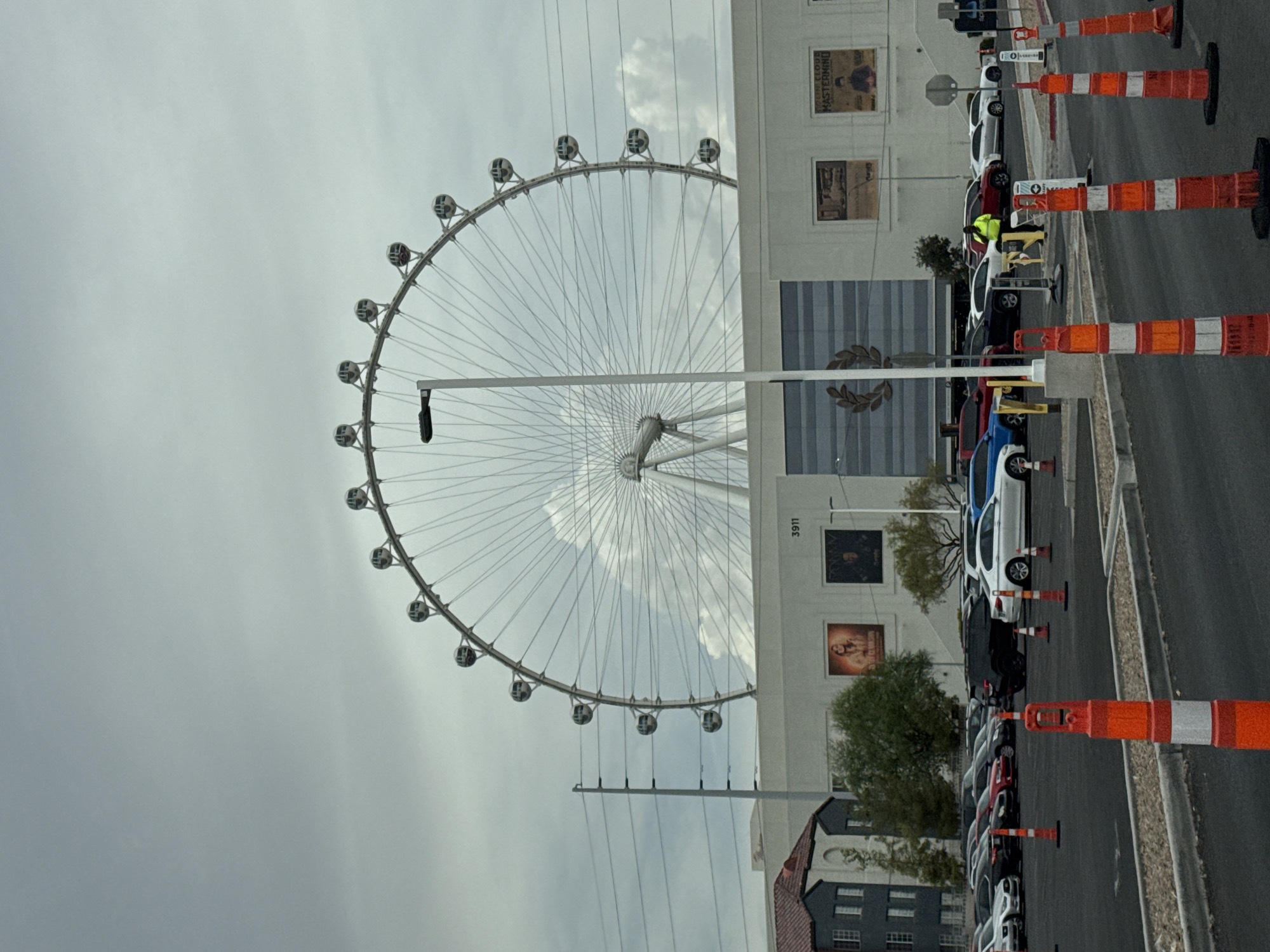 The image shows the High Roller observation wheel in Paradise, Nevada, United States of America, against a cloudy sky. The wheel, which is a large Ferris wheel, dominates the upper portion of the frame. Below the wheel, a modern building with large advertisements on its facade is visible. A parking lot filled with various cars is in the foreground, with several orange traffic cones placed on the asphalt. A lone individual wearing a bright yellow vest is standing in the parking lot. The scene appears to be an outdoor setting during the day.