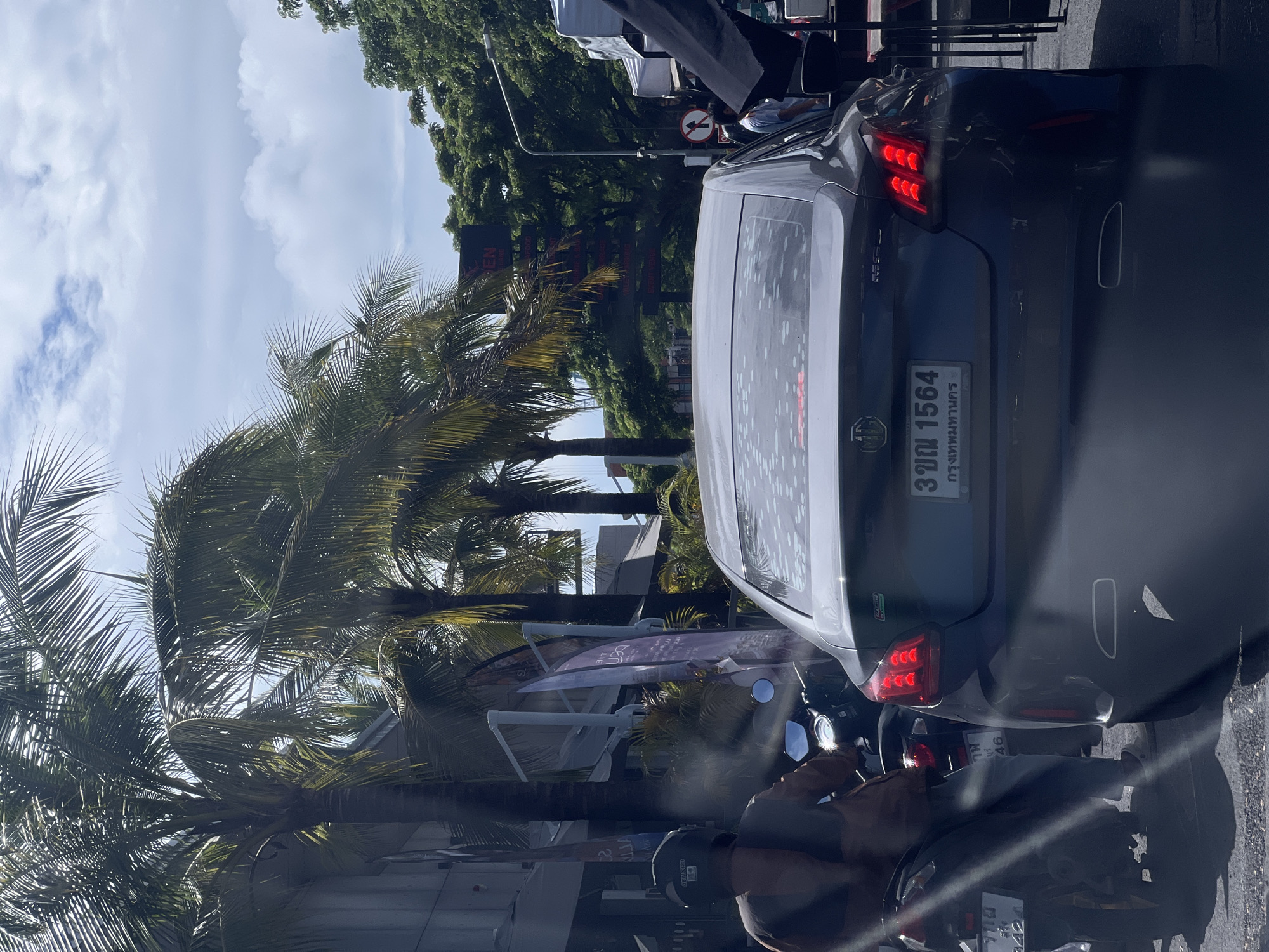 In an outdoor urban scene under a partly cloudy sky, traffic moves along a street lined with palm trees and buildings. A dark-colored sedan is viewed from the rear, its license plate reading "3 ฎม 1564" with Thai characters below. To the left, a person wearing a helmet and a brown jacket rides a scooter, also with a visible license plate. In the background, there are more palm trees, green foliage, and structures, including a sign with red lettering and directional arrows pointing to various establishments. The street appears to be busy with vehicles and pedestrians.