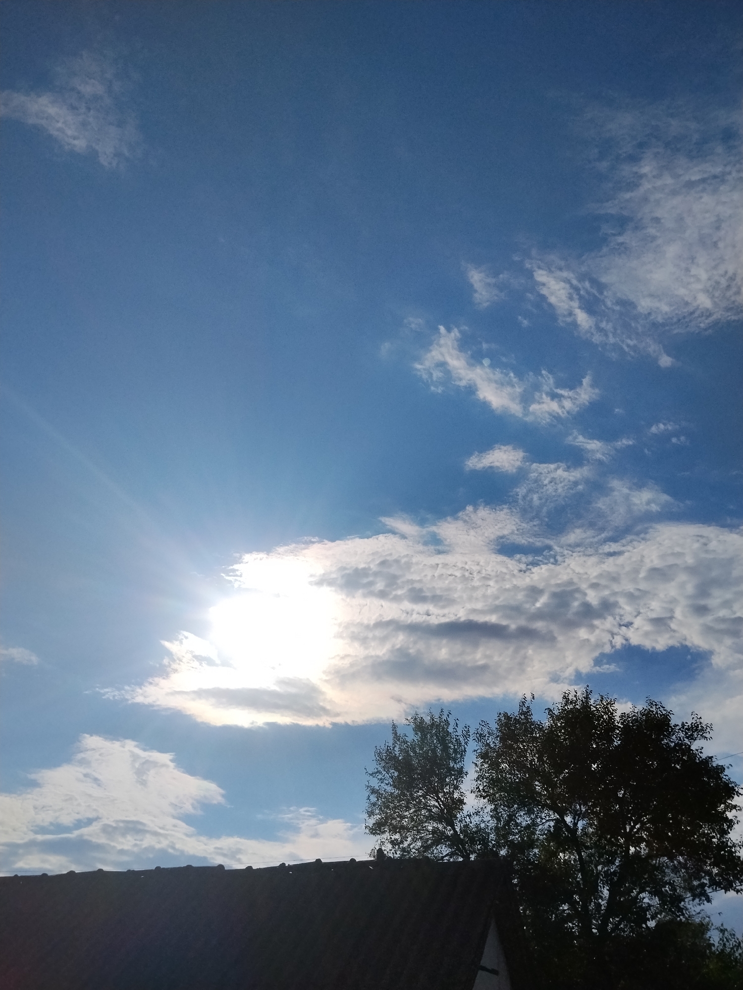 The image depicts a bright, partially cloudy sky over a rooftop and trees, suggesting an outdoor scene in Ustyvytsia, Ukraine. The sun is visible, peeking through a band of clouds on the left side of the frame, casting rays of light into the blue sky. Scattered white clouds are present throughout the upper portion of the image. In the lower section, the dark silhouette of a building's roofline runs horizontally across the frame. To the right of the roofline, two silhouetted trees with dense foliage stand against the sky. The overall impression is a serene view of a partly cloudy day with the sun shining.