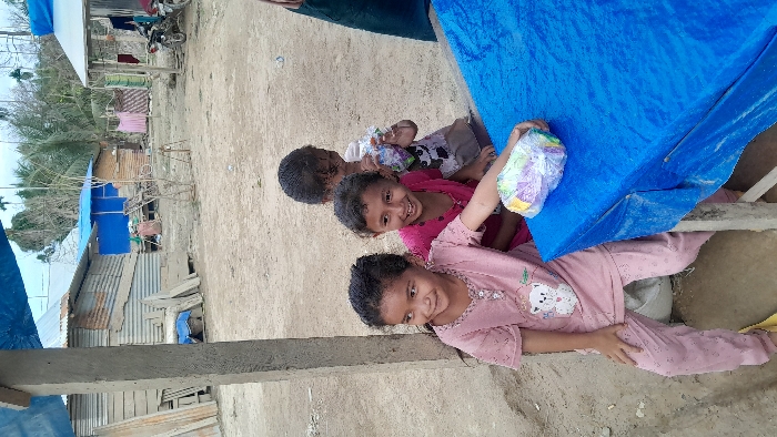 The image depicts an outdoor scene in Rumohrayeuk, Indonesia, centered on three young children near a makeshift table. The ground is dry, light brown earth.
**People:**
A young female child on the left, with dark hair, wears a light pink short-sleeved top featuring a white teddy bear graphic and matching polka-dotted pants. She smiles and looks towards the viewer's left. A second young female child, positioned centrally behind the first, wears a bright pink short-sleeved top, smiles broadly, and also looks to the viewer's left. Her right arm is extended towards the table. A third young child, partially visible behind and to the right of the second child, holds a clear plastic bag filled with multicolored items. This child also looks left with a slight smile. A dark fabric, possibly a garment, is visible on the far right edge, suggesting another person partially out of frame.
**Objects:**
A rudimentary table, likely constructed from wood, is covered with a royal blue tarp or plastic sheet. A clear plastic bag containing colorful items (appearing to be snacks or candies) rests on this blue surface. A rough concrete pillar stands vertically on the left side of the frame. In the background, several single-story structures are visible, constructed from corrugated metal sheets, some showing signs of rust, and wooden planks. At least two of these structures feature blue tarp roofs. A clothesline with drying laundry (pink and white fabrics) is strung between two background structures. Two motorcycles are parked near a structure on the far right of the background. Stacks of wooden planks and other building materials are present near the structures.
**Environment:**
The setting appears to be an informal settlement or rural village area. Sparse trees, including palm trees, are visible in the distant background beneath a bright, possibly overcast sky.