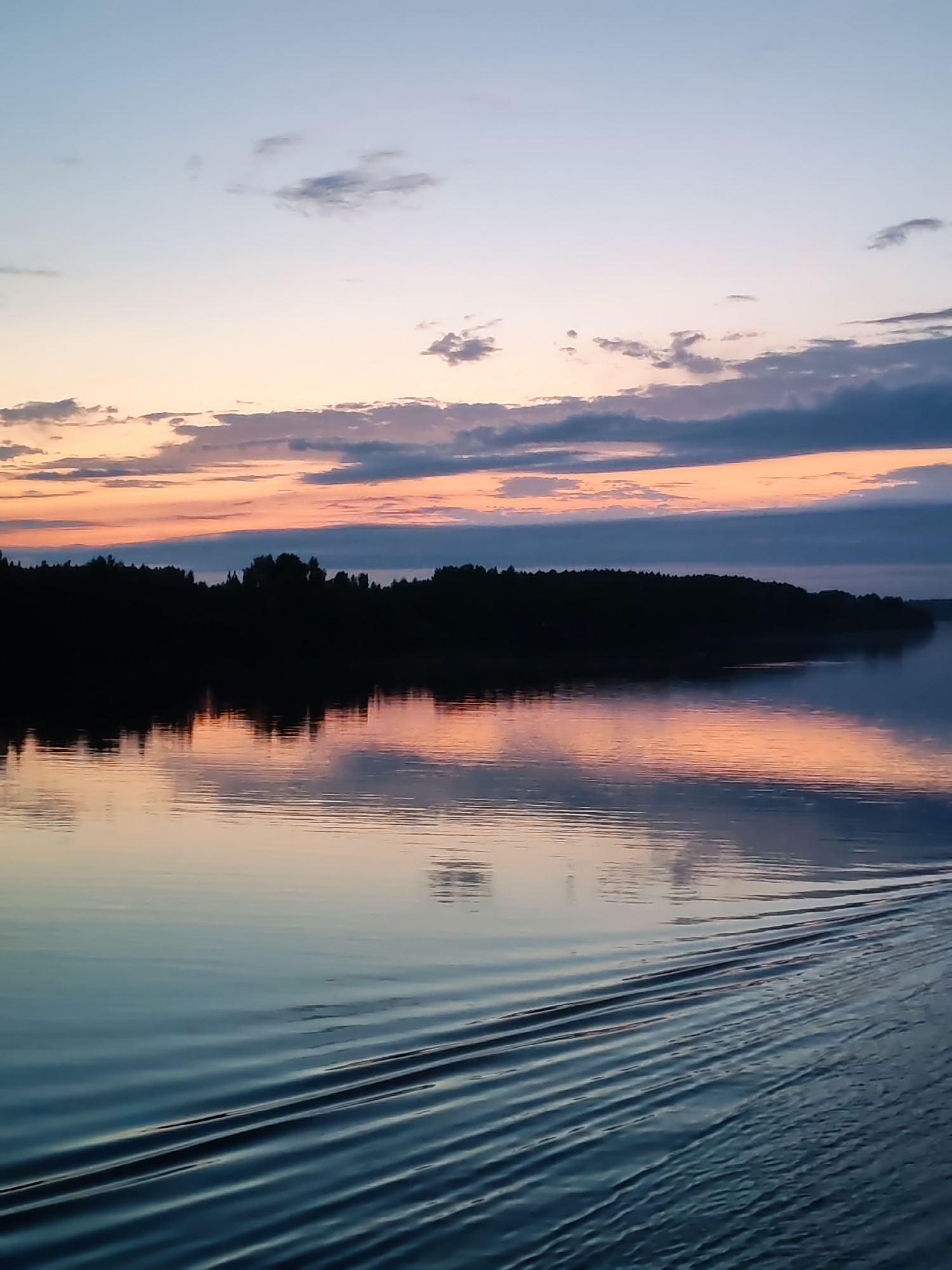 The image depicts a body of water, possibly a lake or wide river, at dusk or dawn. The sky is a mix of light blue, pale orange, and gray clouds. A dark silhouette of trees lines the horizon on the opposite shore. The water reflects the colors of the sky, creating a mirrored effect. Ripples are visible on the water's surface, particularly in the lower portion of the image, indicating movement or waves. The location is unknown.
