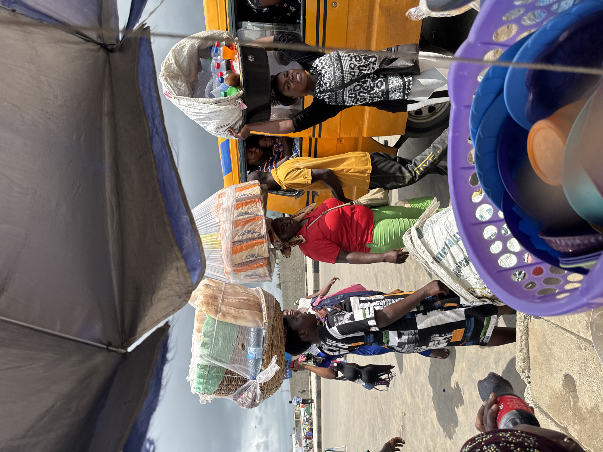 Three women are walking along a road, carrying heavy loads on their heads. The first woman is carrying a basket filled with food, including bread and water. The second woman is carrying a package of baked goods. The third woman is reaching up to a yellow bus with a bag of plastic bottles. Two children are visible inside the bus. One woman is standing on the street near the bus. A partially visible large umbrella shades the women. A purple basket filled with bowls is visible to the right. The location is Ajara, Nigeria, evident from the style of dress and goods being sold.