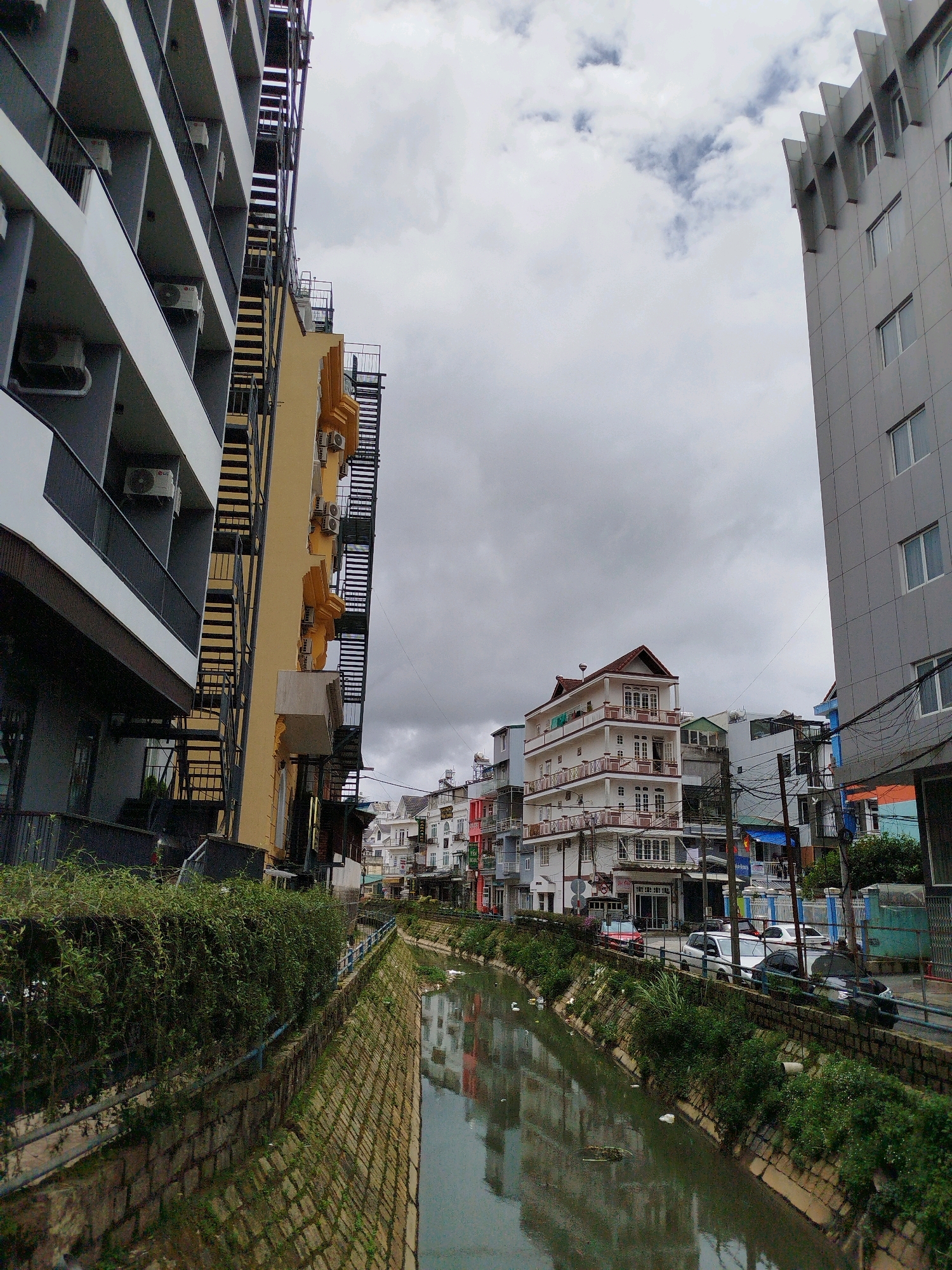 The image depicts a canal flowing through an urban area in Da Lat, Vietnam. The canal, lined with stone walls and overgrown vegetation, reflects the buildings that stand on its banks. These buildings are a mix of modern multi-story structures and older, more colorful houses, creating a varied urban landscape. Fire escapes are visible on some of the buildings, suggesting a dense living environment. Several vehicles, including cars and a small truck, are parked or driving along the road adjacent to the canal. The sky is overcast, with a significant amount of cloud cover. The overall scene conveys a sense of daily urban life in a Southeast Asian city, with the canal serving as a central element in the streetscape.