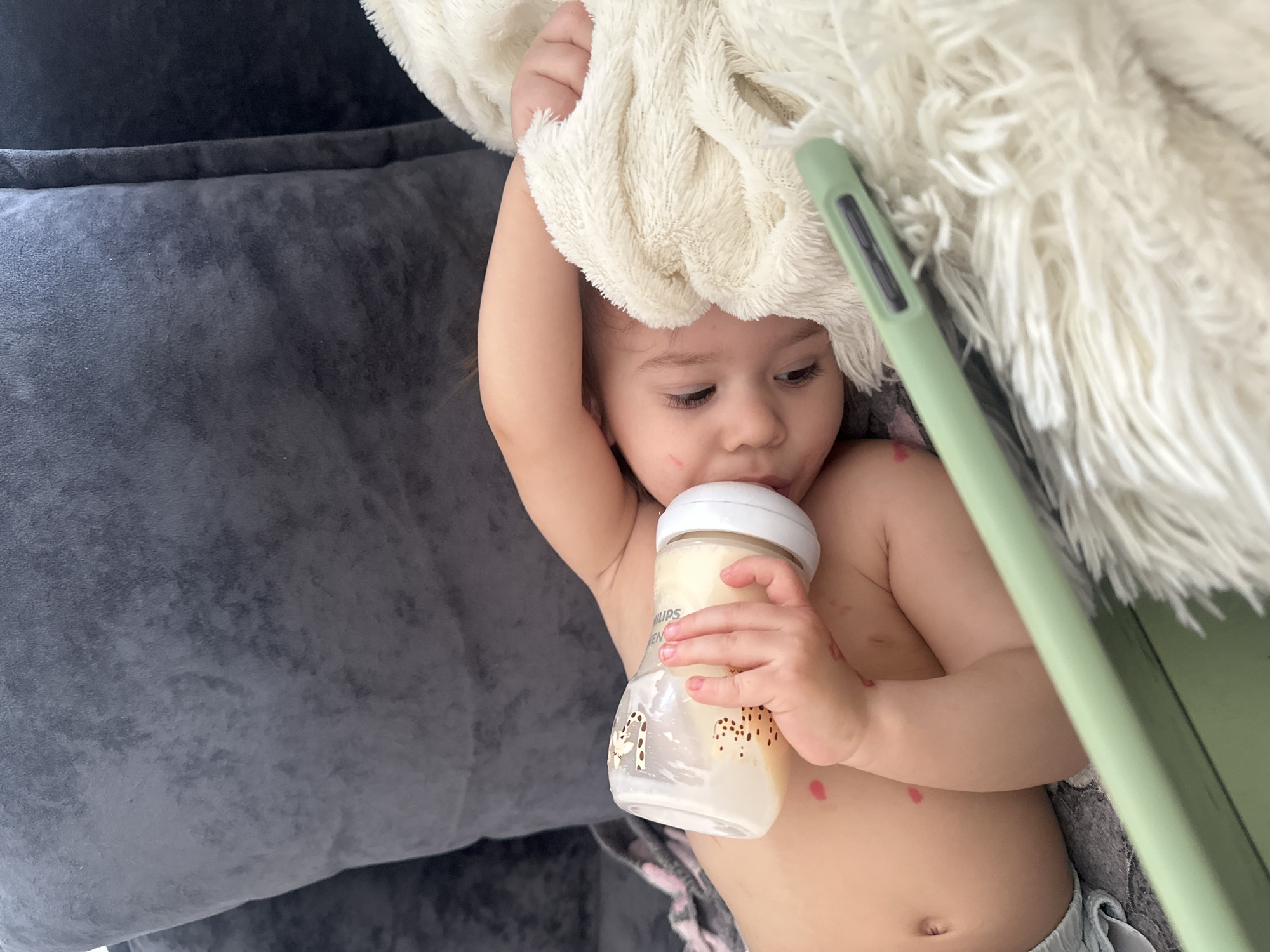 A high-angle, close-up shot captures a baby or toddler lying down, holding a Philips Avent bottle filled with milk and drinking from it. The child, with fair skin and light brown hair, is partially covered by a soft, fluffy white blanket draped over their head and upper body. Their gaze is directed downwards and to the right, indicating focus on the bottle. The child's chest and right arm are visible, showing several small, reddish spots, and a faint red scratch mark is present on their right cheek. They are wearing light-colored, patterned shorts or a diaper.
The child is resting on a dark gray, textured surface, likely a sofa or a bed. To the upper right of the frame, a light green rectangular object, possibly a tablet or smartphone encased in a cover, is partially visible, positioned near the child's head and partially beneath the white blanket. The white blanket has a shaggy texture, providing a soft backdrop to the child.
The only visible text is "PHILIPS AVENT" printed vertically on the side of the baby bottle, which also features a subtle giraffe pattern. The image conveys a calm, intimate moment, likely indoors during the daytime. This scene is located in Kharnavy, Russia.