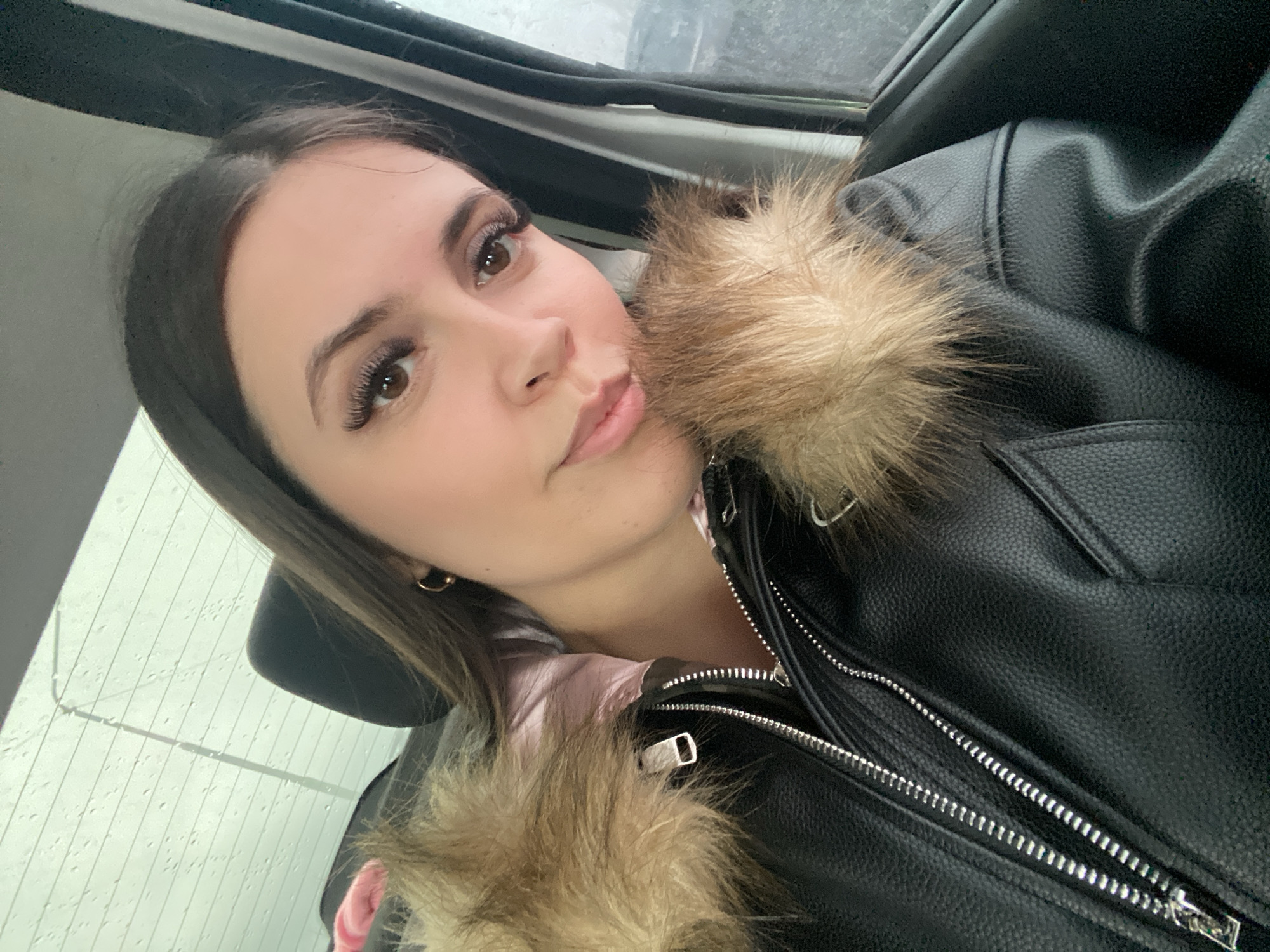 A person with long brown hair is taking a selfie, wearing a black leather jacket with fur trim, inside a vehicle. The backdrop shows rain on a window, which suggests the environment may be outdoors. No information is present to confirm that the location is in the city of Chaikovsky, Russia.