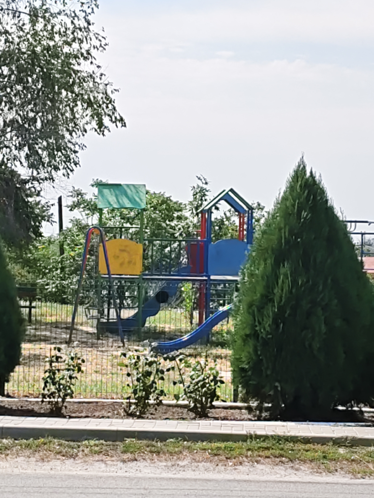 The image shows a children's playground in Pukhlyakovskiy, Russia. The playground features a blue and red slide, a yellow swing seat, and a green canopy. The playground is enclosed by a metal fence, and small green bushes are planted in front of it. Two large green trees are visible on either side of the playground, and a large tree with green leaves is in the background. The sky is clear and blue with some clouds. The ground is covered with dirt and gravel, and there is a paved walkway in the foreground.