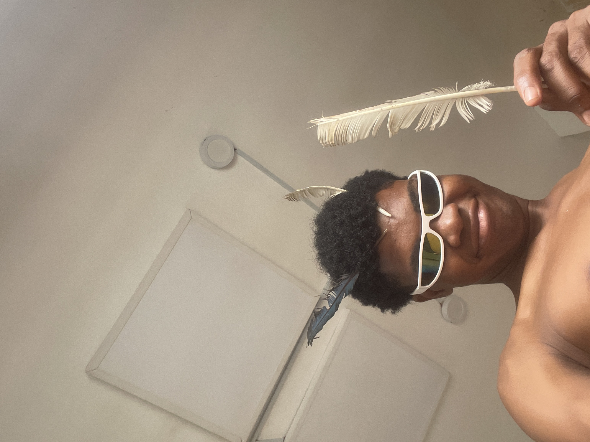 The image shows a person wearing white-rimmed sunglasses and holding a feather. The person has dark, curly hair with two feathers sticking out of it. The background appears to be a ceiling with two large square panels and a round light fixture. The image is a close-up shot, taken from a low angle, with the person looking up towards the camera. There is no discernible information about the location in the city of Ado, Nigeria within the provided image.