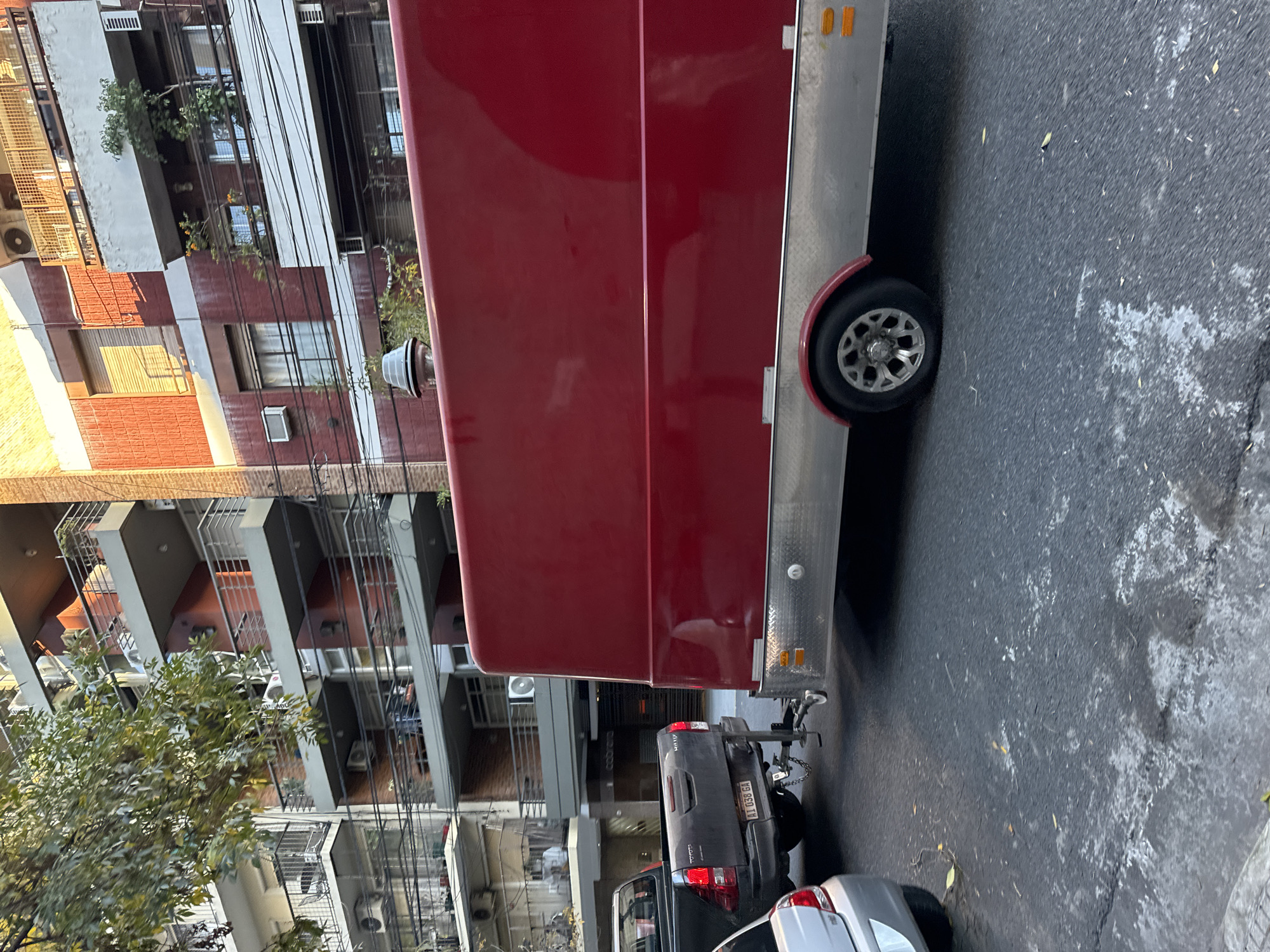 The image shows a street scene in Buenos Aires, Argentina. A red food trailer is parked on the right side of the street, attached to a dark gray Toyota Hilux pickup truck. A silver car is parked behind the truck. The buildings lining the left side of the street are a mix of brick and concrete apartment blocks, typical of the urban architecture in Buenos Aires. Trees are visible, partially obscuring the lower floors of the buildings. Power lines crisscross the sky in front of the buildings. The street surface is asphalt with some visible wear. The license plate on the truck reads "AI 038 GA". The overall lighting suggests it is daytime. There are no people visible in the frame.