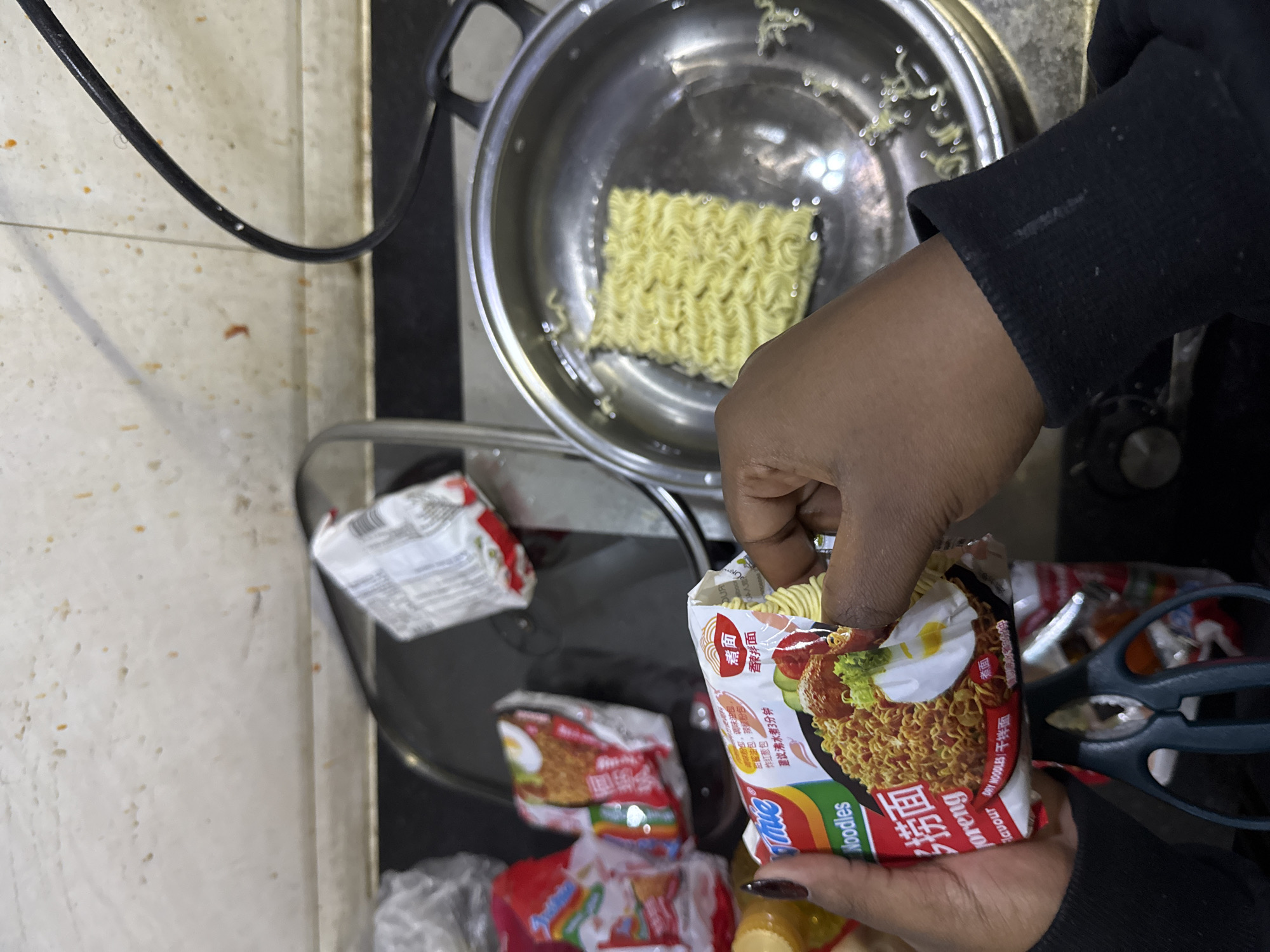 A person is preparing instant noodles. A block of dried noodles sits in a stainless steel pot filled with water. The person, wearing a black long-sleeved shirt, is reaching into an open Indomie Mi Goreng instant noodle package. Additional unopened packages of the same noodles are visible next to a pair of scissors. The scene takes place on a countertop with a mottled pattern. While the noodle package indicates "DRY WOODLES/干拌面", the scene does not depict the city of Zhongtun, China.