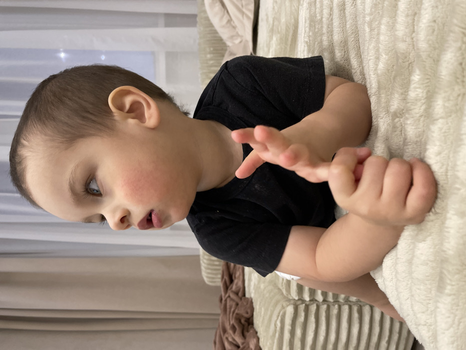 A young child, appearing to be a toddler, is situated in an indoor residential setting. The child has fair skin, short light-brown hair, and blue eyes, with a noticeable reddish flush on both cheeks. They are dressed in a black short-sleeved t-shirt, with a white undergarment, possibly a diaper, visible beneath. The child lies prone on a light-colored, horizontally ridged upholstered surface, consistent with a bed or sofa. Their head is turned sharply to the left, with their gaze directed upwards and to the left. The child's left hand is fisted and rests on the upholstered surface, while the right hand is held with fingers partially splayed.
In the background to the left, a folded brown textured blanket is visible on an adjacent upholstered section. Further behind, a window is covered by both translucent white sheer curtains and heavier beige drapes. A faint, diffused light source is discernible through the sheer curtain on the right side of the window. The specific location within the city of Izhevsk, Russia, is not identifiable from the interior details depicted in the image. No other individuals are present or interacting with the child within the visible frame.