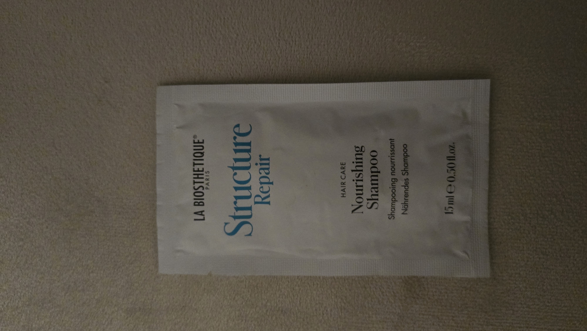 A small, white, foil packet lies flat on a textured, beige surface. The packet is oriented vertically and is centered within the frame. The front of the packet features text printed in various fonts and colors. At the top, "LA BIOSTHETIQUE" is printed in black, followed by "PARIS" in smaller black font. Below this, "Structure Repair" is written in a bold, blue font. Further down, "HAIR CARE" is printed in small black text. Beneath that, "Nourishing Shampoo" is displayed in a prominent black font. Finally, at the bottom, the text "15ml e 0.50 fl.oz." is printed in black. The beige surface appears to be a textile, possibly a towel or a soft blanket, with a subtle, repeating pattern. There is no indication of the location being in Batumi, Georgia, or any other specific city. There are no people, actions, or interactions visible in the image.