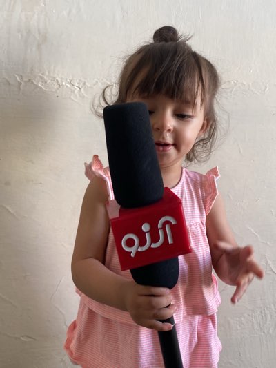 A young child, likely a toddler, is captured in a close-up, waist-up shot, holding a microphone. The child has dark hair, partially pulled into a small bun on top, with bangs framing their face. They are wearing a light-colored, possibly pink or peach, striped top with ruffled cap sleeves. The child's expression is one of curiosity and engagement, with a slightly parted mouth as if speaking or concentrating.
The central object is a large, black microphone, featuring a prominent red, square-shaped microphone flag. The flag displays white text in Arabic or Persian script, which reads "آرزو" (Arzo or Arezoo), meaning "wish" or "hope." The child holds the microphone with both hands, positioned centrally in front of their chest and face, suggesting they are playing or mimicking a reporter.
The background is a plain, light-colored wall, possibly off-white or beige, showing some texture and minor irregularities. The setting appears to be indoors, with soft, even lighting, indicative of daytime. The overall scene suggests a moment of innocent play and imagination. This image offers a glimpse into a child's life in Kandahar, Afghanistan, engaging in imaginative role-play.