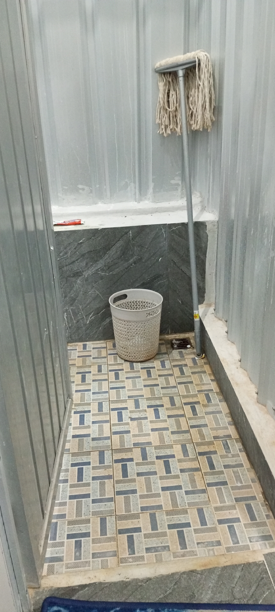 The image shows a utility area, possibly a restroom or cleaning closet, with a moped leaning against a corrugated metal wall. A white woven basket is on the floor, and the floor is tiled with a pattern of beige, blue, and grey rectangles. A drain is visible on the floor near the basket. A red object, possibly a cleaning supply, is on a ledge above the basket. The overall impression is one of a functional, utilitarian space. The scene is not specific to Parigi, Indonesia.