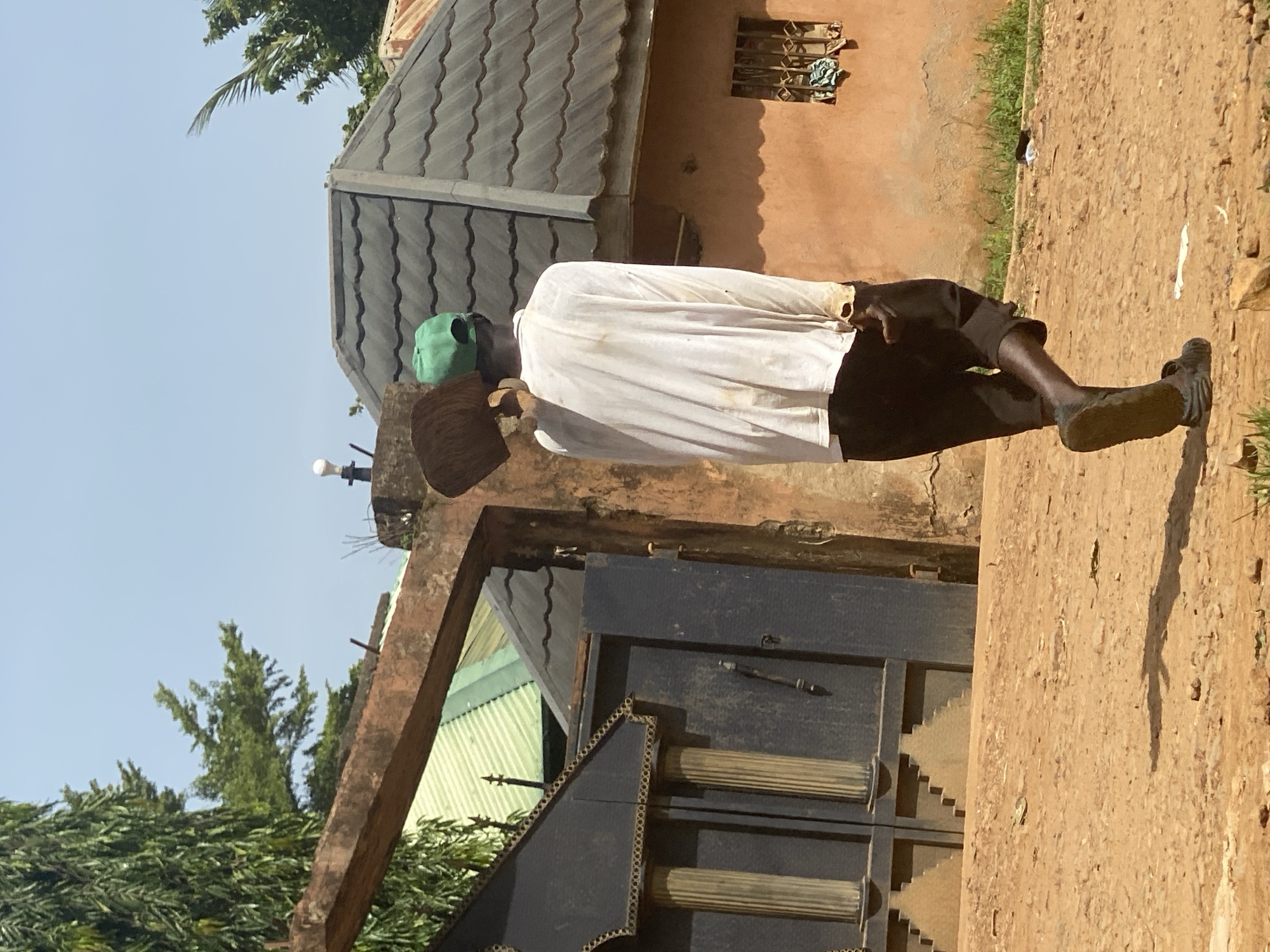A full body shot of an older man, likely indigenous to Maigizo, Nigeria, stands outside a weathered building. He wears a green cap, a light-colored shirt, and dark shorts. He holds an object in his hands. The building features a grey tile roof, peach-colored walls, and a black, ornate gate. The ground is a light brown color. Trees are visible in the background under a clear, blue sky.