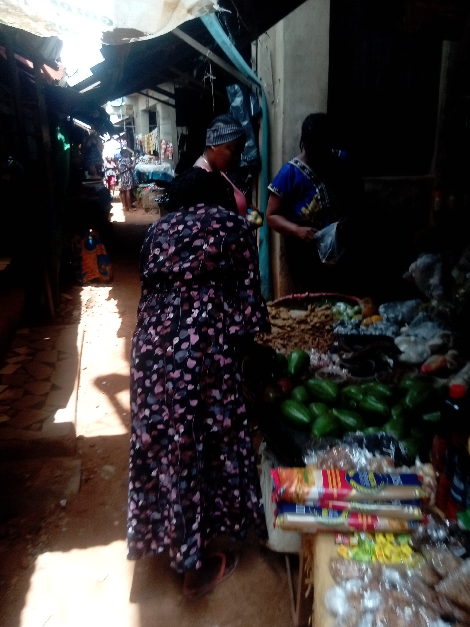 The image depicts a market scene in Ogidi, Nigeria. An individual wearing a floral-patterned dress faces a market stall laden with produce. Green peppers and packages of pasta are visible on the stall. Two other women are also present: one wearing a patterned head covering who appears to be examining items at the stall, and another woman wearing a blue and gold patterned shirt. The market appears to be in a covered, shaded area with other people visible further down a walkway. The scene is illuminated by a combination of natural sunlight filtering through the market structure and darker areas.