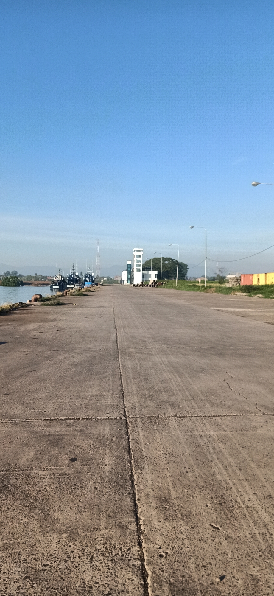 The image shows a clear day at a port in Keudekruenggeukueh, Indonesia. A wide, paved road dominates the foreground, stretching towards a body of water on the left. Several dark-colored boats are docked along the water's edge. In the middle ground, a white building with a tower stands out, accompanied by a radio tower and some large pipes. Green grass lines the sides of the road, and in the distance, hills are visible under a bright blue sky. There are no people in the image.