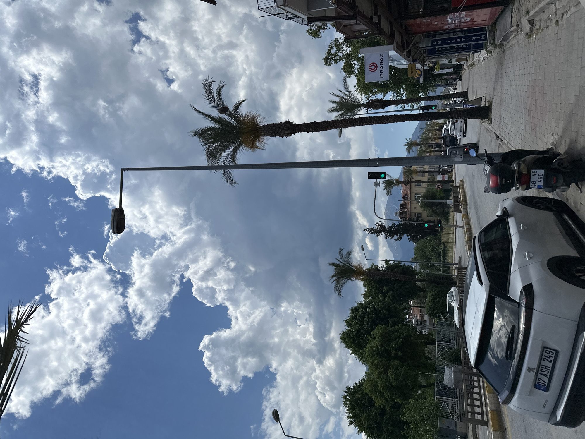 The image shows a street scene in Fethiye, Turkey. The camera is at ground level looking down the street, which is lined with buildings on the left and parked vehicles on the right. The sky is partly cloudy, with large white clouds and patches of blue. A tall palm tree stretches up on the left side of the street, near a street light. Further down the street, there are more palm trees, traffic lights showing green, and buildings, with mountains visible in the distance. A motorcycle is parked on the right side of the street next to a white car. The license plate of the white car reads "07 ASH 249." A sign for "IPRAGAZ" is visible on the left side of the street. The street pavement is made of interlocking bricks.