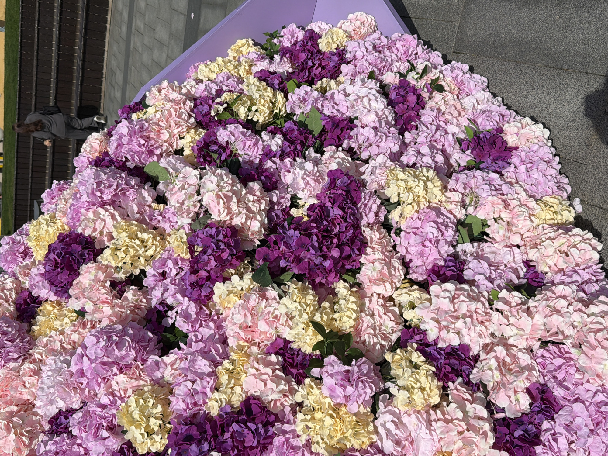 This image showcases a large, vibrant display of artificial flowers, primarily hydrangeas, arranged in a dense, mound-like formation. The dominant colors of the flowers are various shades of purple (from light lavender to deep violet), pink, and pale yellow or cream. Small green leaves are interspersed among the numerous blooms. The floral arrangement appears to be set against a light purple frame or backing, visible on the upper left and right edges.
In the upper-left background, a person with long, dark hair, dressed in a grey jacket and dark trousers, is partially visible, standing near a dark, tiered structure that could be seating or steps. Adjacent to this structure, a strip of green grass is visible. The foreground and right side of the image show grey stone paving, suggesting an outdoor, urban setting.
The scene is brightly lit by what appears to be strong sunlight, indicating daytime and clear weather. The image captures a static display rather than an ongoing activity, creating a decorative and colorful atmosphere. No text is visible in the image. Given the provided context, the location is Moscow, Russia.