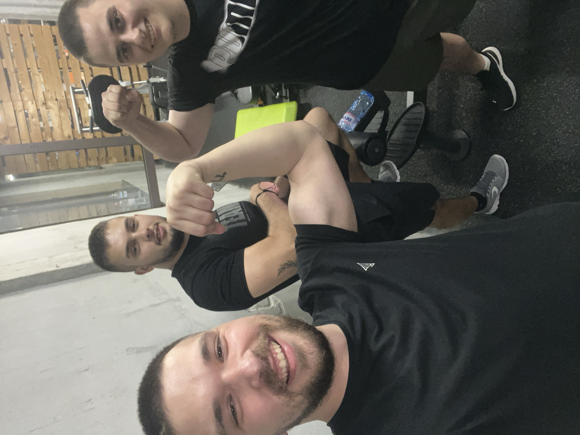 Three men are posing in what appears to be a gym setting. The topmost man in the frame is holding a dumbbell and flexing his bicep. The man in the middle is also flexing his bicep. The man at the bottom of the frame is smiling directly into the camera. All three men are wearing dark-colored shirts.
A leg press machine is visible in the background behind the men, along with a water bottle. The flooring has a speckled pattern. There are no distinct details visible to confirm that this picture was taken in Sofia, Bulgaria.