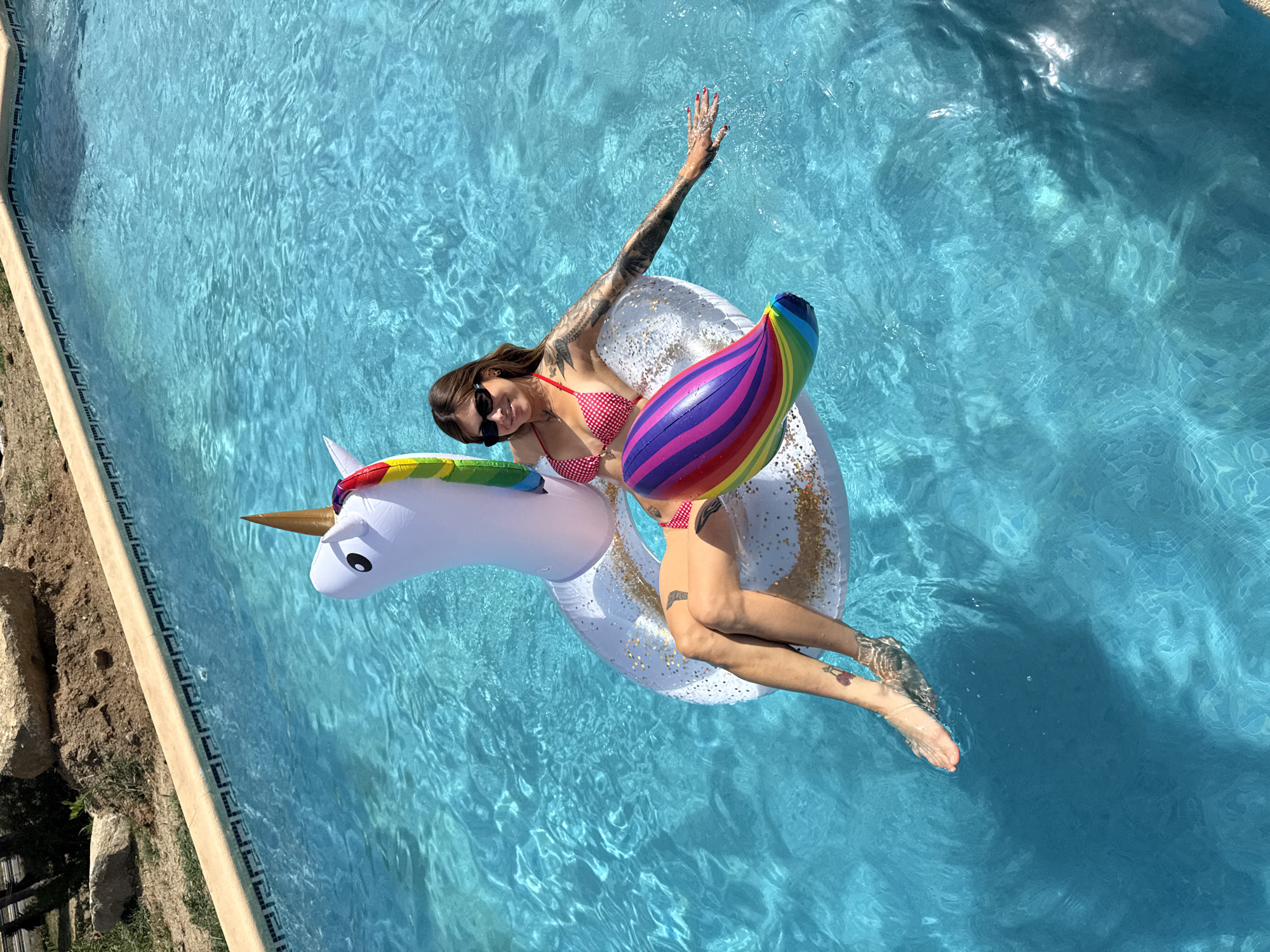 A woman wearing a red and white polka dot bikini and sunglasses floats on an inflatable unicorn in a swimming pool. The pool water is a vibrant blue, with sunlight creating patterns on the surface. The woman is positioned on the unicorn's body, with her legs extended and partially submerged in the water. She has tattoos on her left arm and leg. The unicorn float is white with gold glitter accents, a rainbow-colored mane, and a golden horn. In the background, the edge of the swimming pool is visible, constructed with light-colored coping stones and patterned tiles. Beyond the pool, there is a dry, earthy area with some rocks and sparse vegetation, suggesting an outdoor setting in a dry climate. This scene is located at a private residence with a swimming pool in Sant Vicenç de Montalt, Spain.