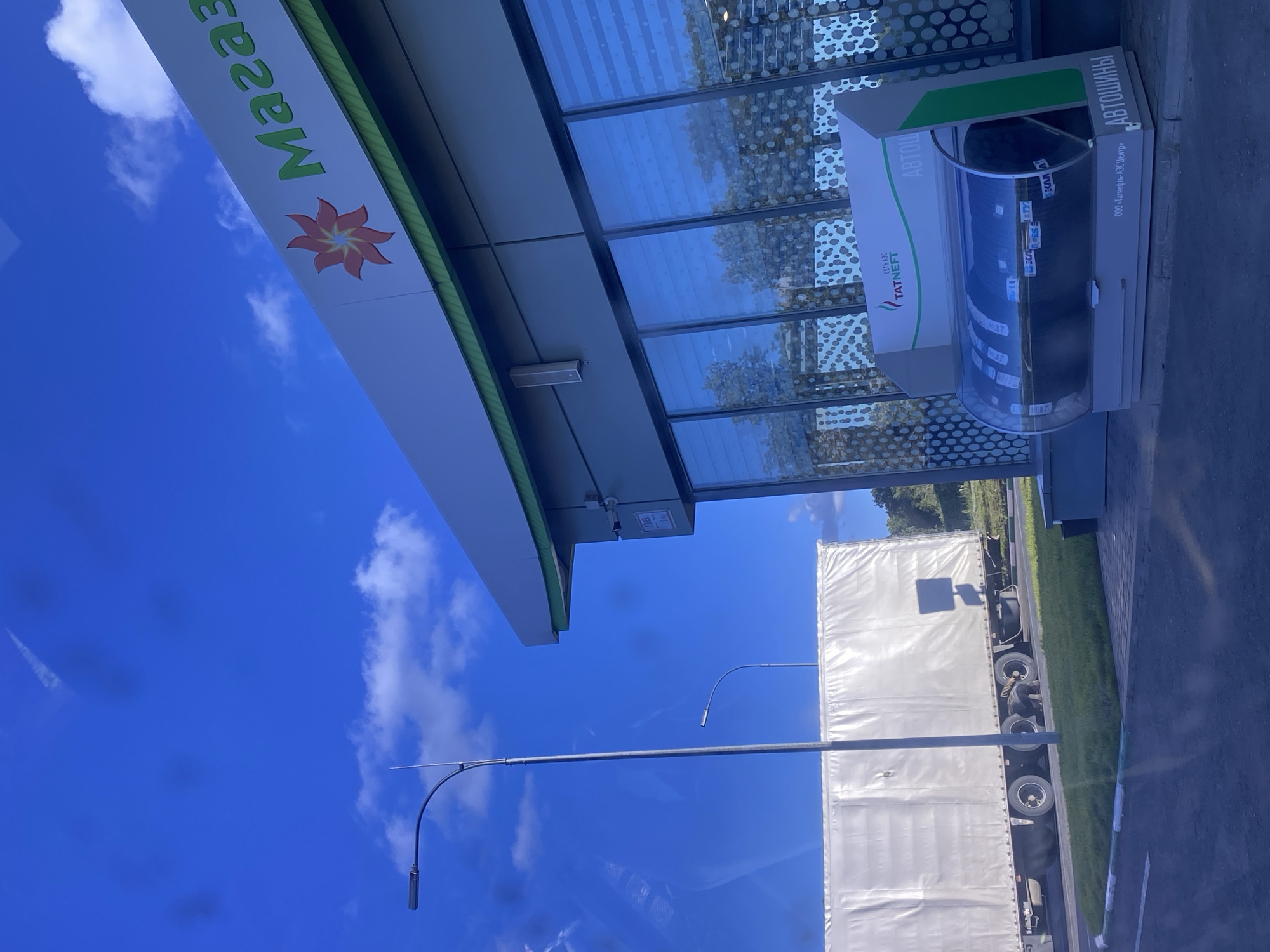 The image depicts a gas station with a "Magaza" sign and a logo featuring a red flower. In the foreground, a display unit containing car tires is visible. To the left, a large white truck trailer is parked on the side of the road. A street lamp stands prominently in the mid-ground. The background shows trees and a bright blue sky with some clouds. The scene is likely located in a suburban or rural area, given the presence of the truck and open space. The specific location in Заглядовка, Russia, is inferred from external information, as there are no explicit visual cues in the image to confirm this.
