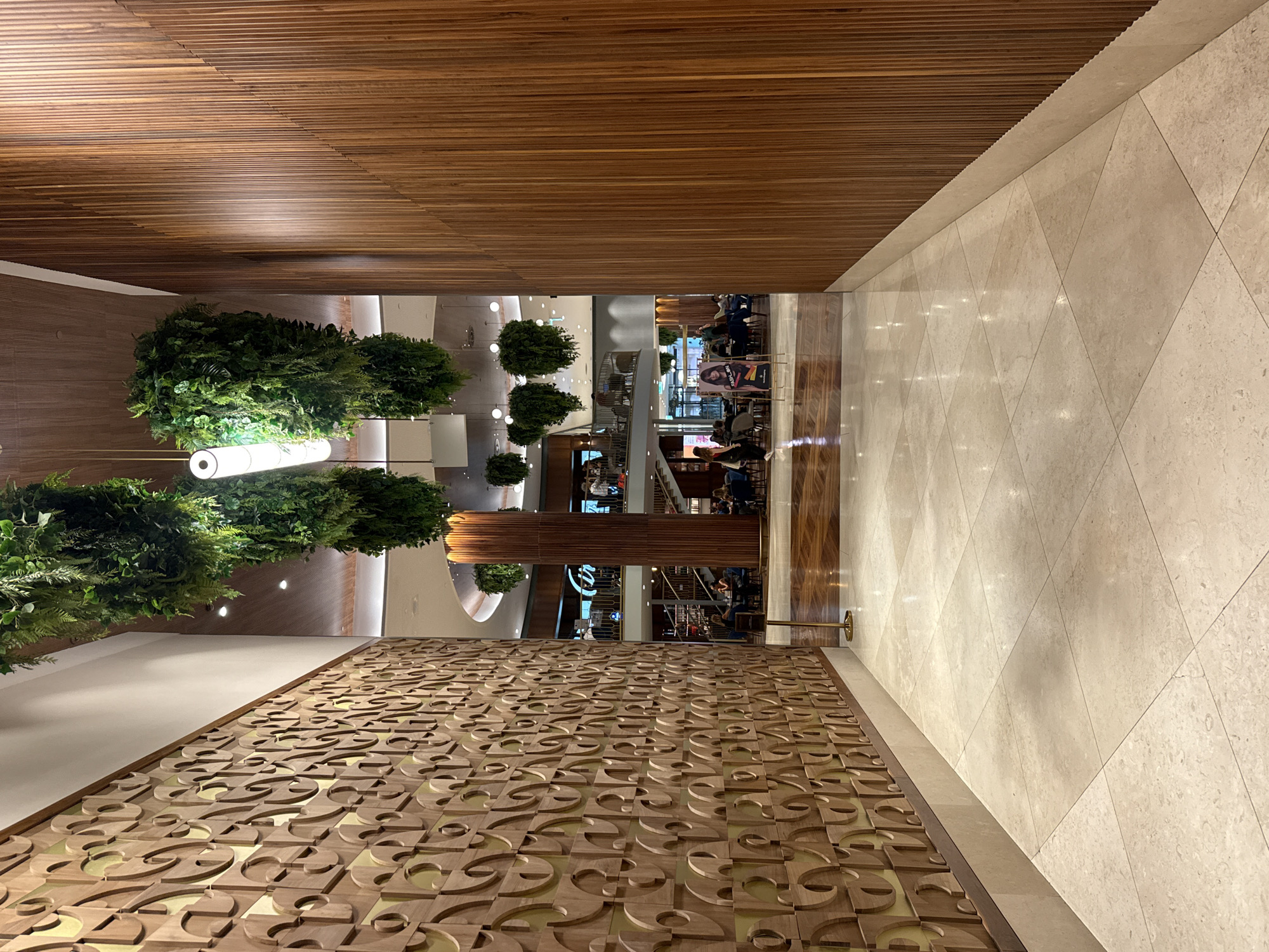 The image shows an interior shot of what appears to be a modern, upscale shopping mall or commercial establishment, possibly located in Matosinhos, Portugal. The scene features a long, wide hallway with two distinct textured walls. The wall on the left is adorned with a decorative wooden panel featuring a repeating, intricate pattern of curved, interconnected shapes, resembling stylized letters or abstract designs. This wall has a light-colored baseboard and extends upwards to a white ceiling or upper wall.
The wall on the right is constructed of vertical wooden slats, creating a textured, ribbed effect that runs from floor to ceiling. A light-colored baseboard separates the wooden wall from the floor. The floor itself is made of large, light-colored square tiles, some of which reflect the overhead lighting, creating a polished and slightly glossy appearance.
Hanging from the ceiling are large, cascading arrangements of artificial greenery, adding a touch of organic design to the otherwise modern architecture. Two elongated, cylindrical light fixtures are visible among the hanging plants.
In the background, the hallway opens up to a more public area with multiple levels. People are visible seated at tables in what appears to be a dining area or cafe. There are escalators and staircases, suggesting multiple floors. A large, cylindrical wooden column also stands in the background, adorned with more hanging greenery. A poster with an image of a woman and text that includes "FASHION" is placed near the entrance to the seating area. The overall ambiance is clean, contemporary, and well-lit.