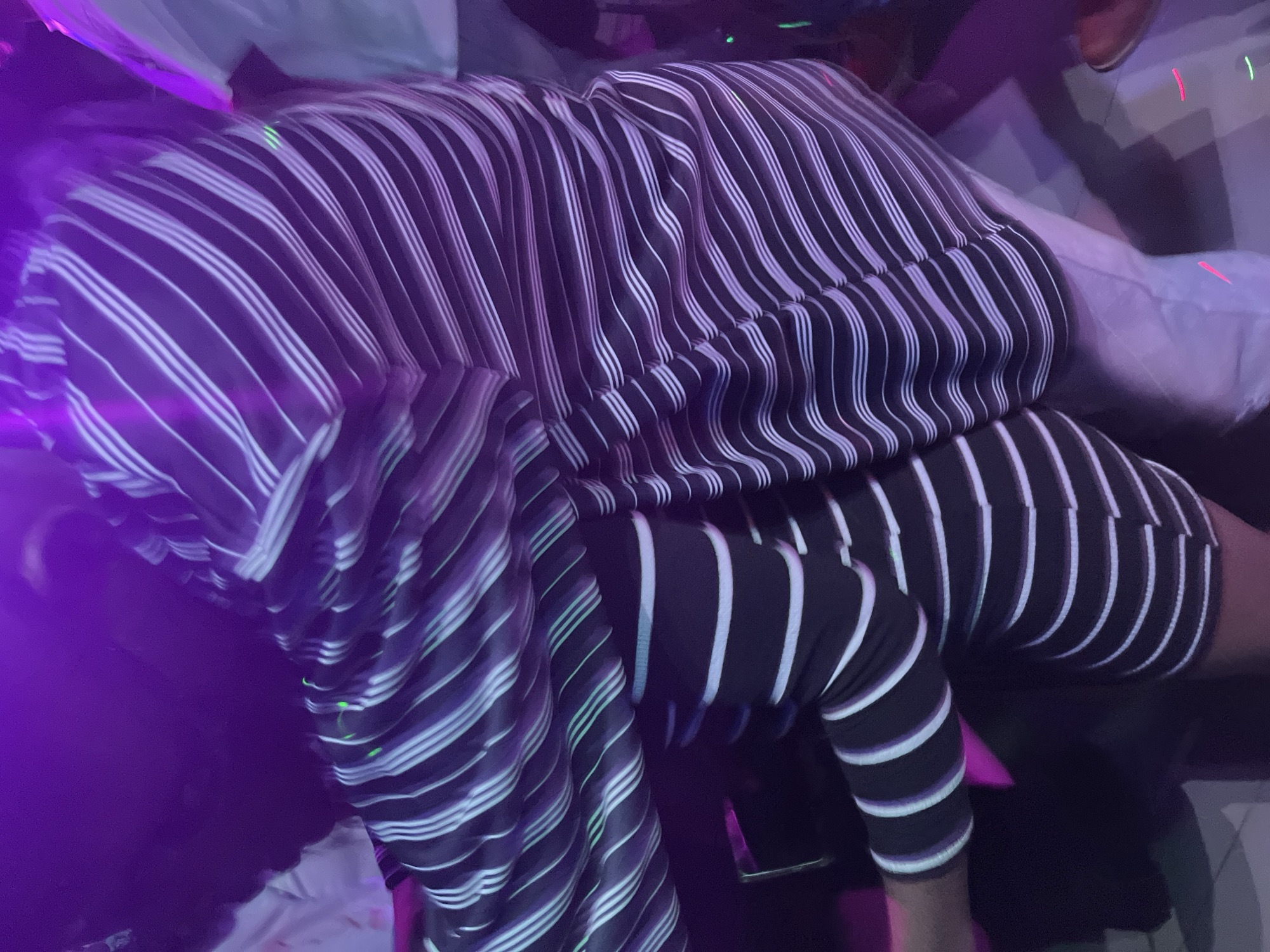 The image depicts a close-up view of two individuals in what appears to be a dimly lit, possibly nightclub or party environment, indicated by the prevalent purple lighting and blurred motion. One individual is positioned behind the other, with their torsos facing each other. The person in the foreground is wearing a black and white striped garment that appears to be a dress or top and skirt combination. The stripes are horizontal and run across the body, with some white and faint green or blue reflective stripes. The person in the background also appears to be wearing a striped garment, possibly a shirt, with similar black and white diagonal stripes. The lower half of the background individual's legs are visible, wearing light-colored pants. The overall scene is dynamic and suggests movement, possibly dancing, due to the blur. The environment is indistinct, with splashes of purple and some areas of darkness. There are no clear identifiable landmarks or context to suggest a specific location within a city.