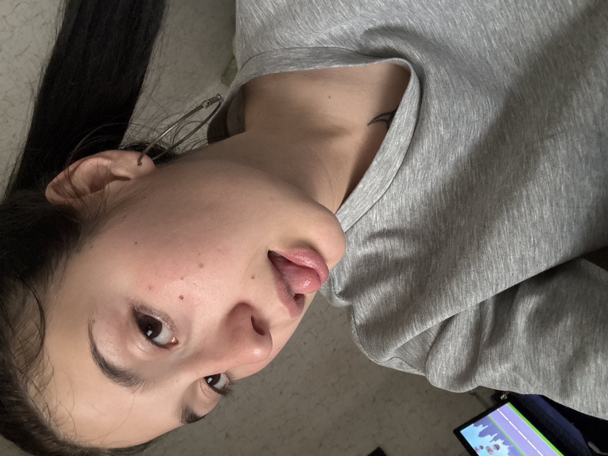 The media file is a close-up, vertically oriented photograph of a person's face. The subject is a young person with dark hair pulled back, revealing their left ear with a hoop earring. They are wearing a grey sweatshirt. Their eyes are open, and their tongue is sticking out slightly. The lighting appears to be indoor, with some slight shadowing. There is a tattoo visible on the person's neck. The background is a textured surface, possibly a wall or fabric. In the lower right corner, a small portion of a screen displaying a video game is visible. There is no discernible text in the image. The image appears to be taken indoors, possibly in a casual setting.