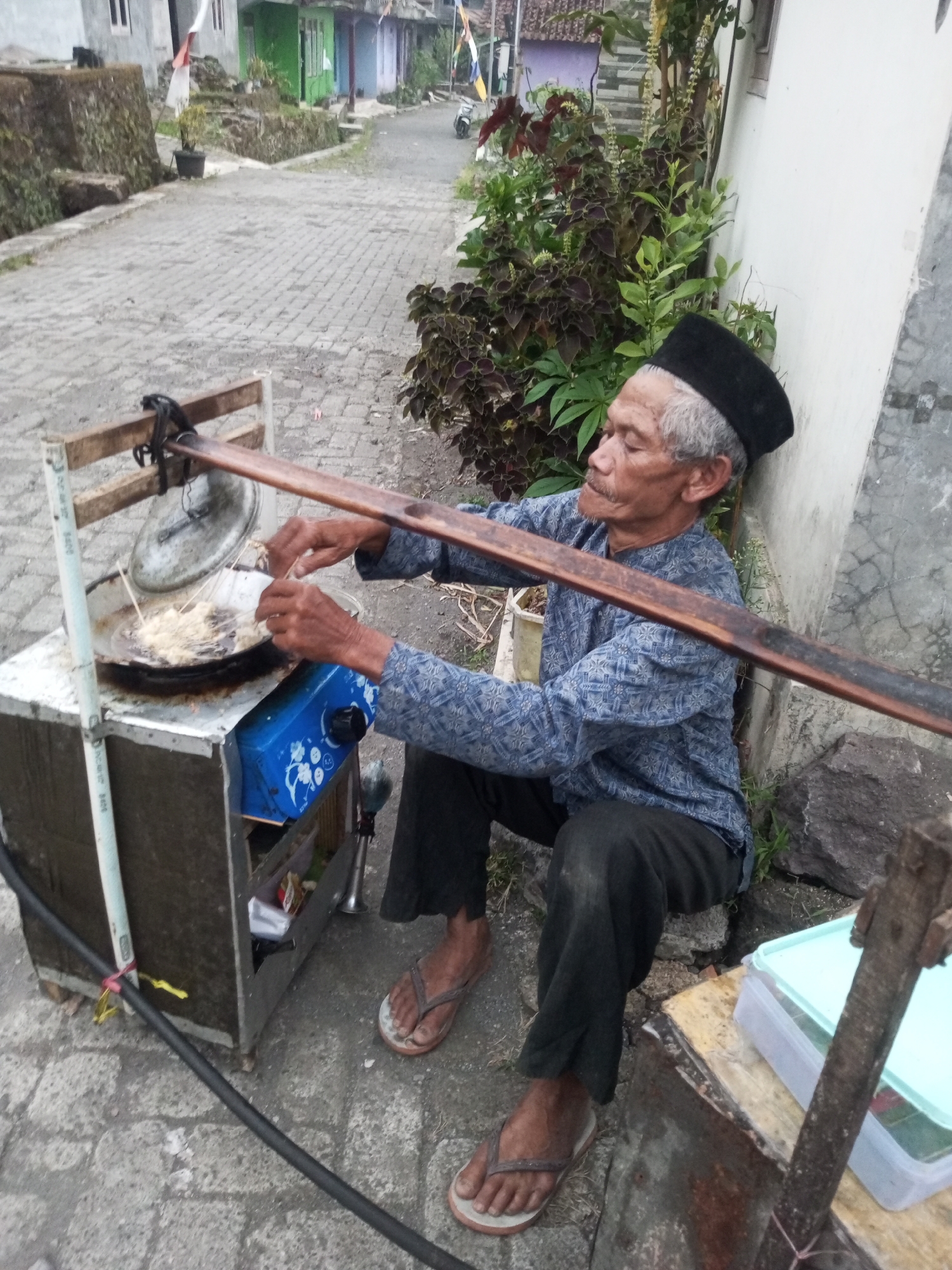In Andongsili, Indonesia, an elderly Indonesian man with gray hair and a mustache, wearing a black peci hat and a blue patterned long-sleeved shirt, is frying food. He is crouched next to a mobile food cart. The cart has a silver exterior, a blue gas stove on the lower shelf, and a pan of hot oil on the top shelf. The man is using skewers to fry small, breaded pieces of food in the hot oil. He is wearing dark pants and flip-flops. The cart is situated on a paved road with stone walls and buildings in the background. Lush green plants are growing along the side of a white building next to the man. The overall scene depicts a street food vendor preparing food outdoors.